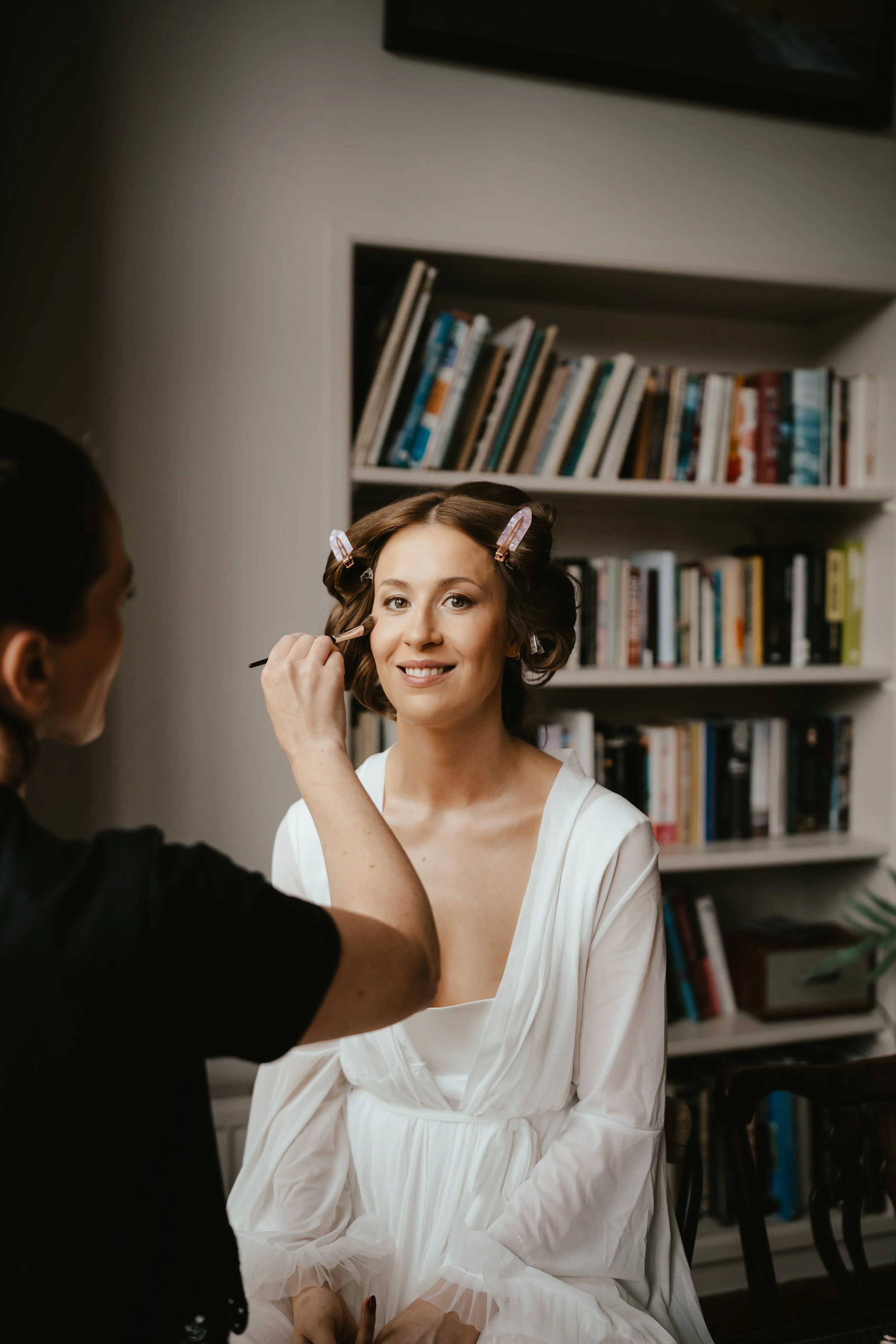 Woman in a white dress with hair rollers getting her makeup done by a makeup artist.