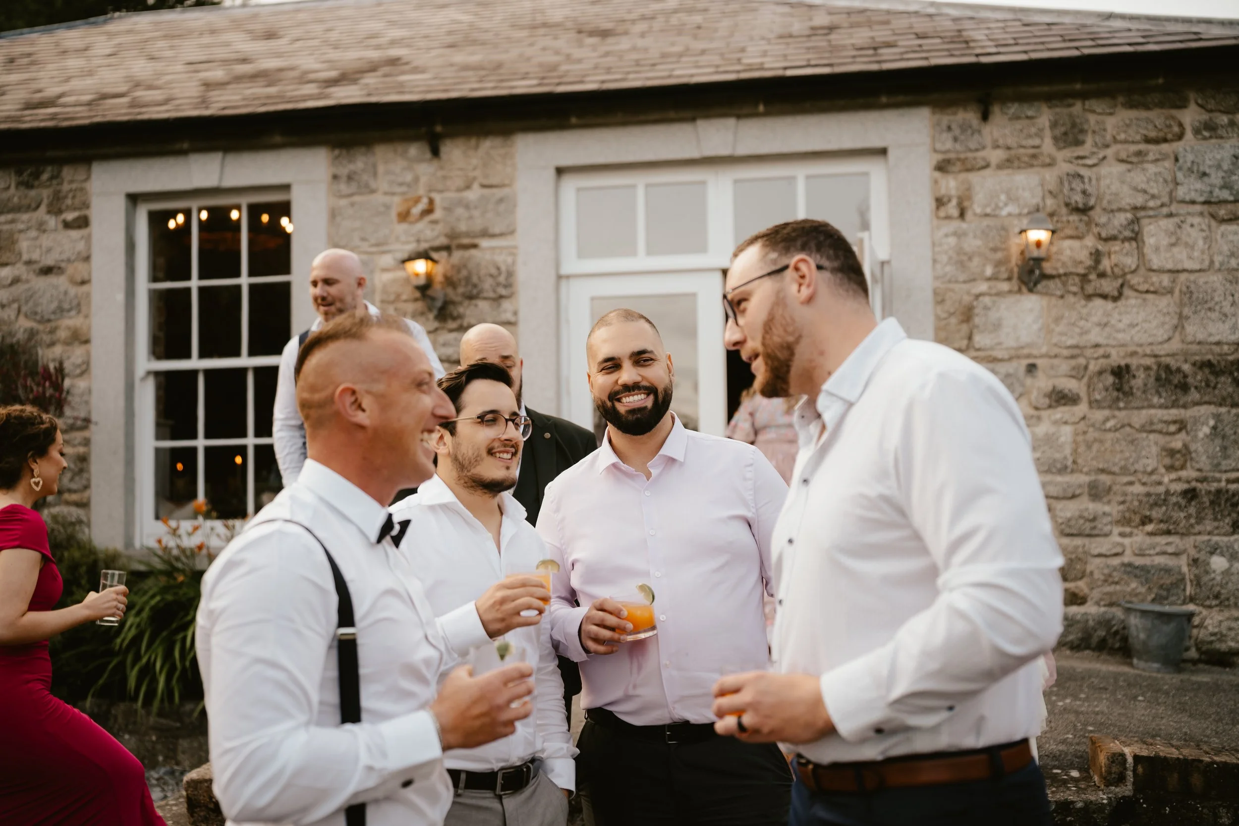 Group of men and women at an outdoor social gathering, enjoying drinks in front of a stone building with lit wall-mounted lights.
