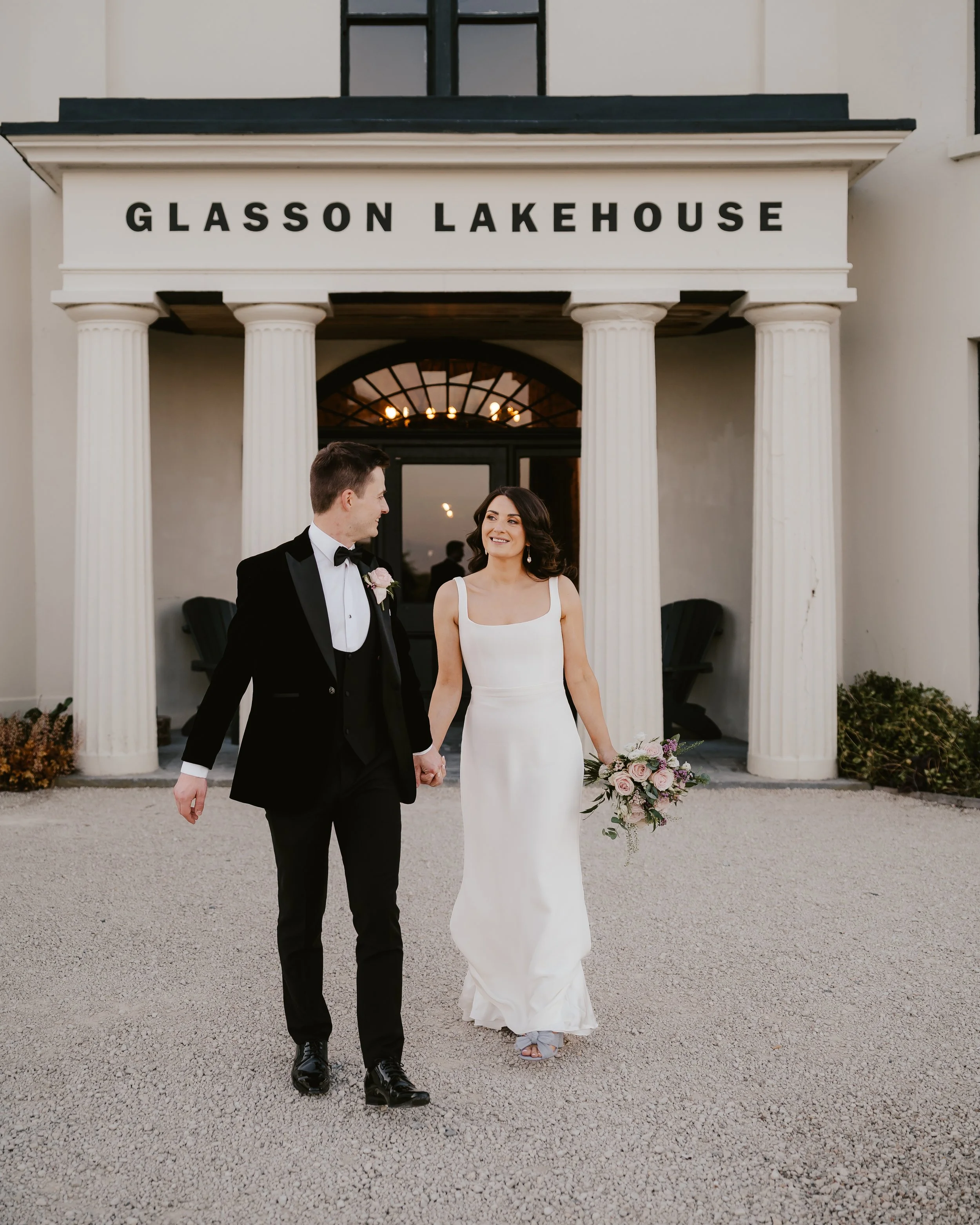 A bride and groom holding hands and walking in front of the Glasson Lakehouse, a wedding venue with classical columns and a sign above the entrance.