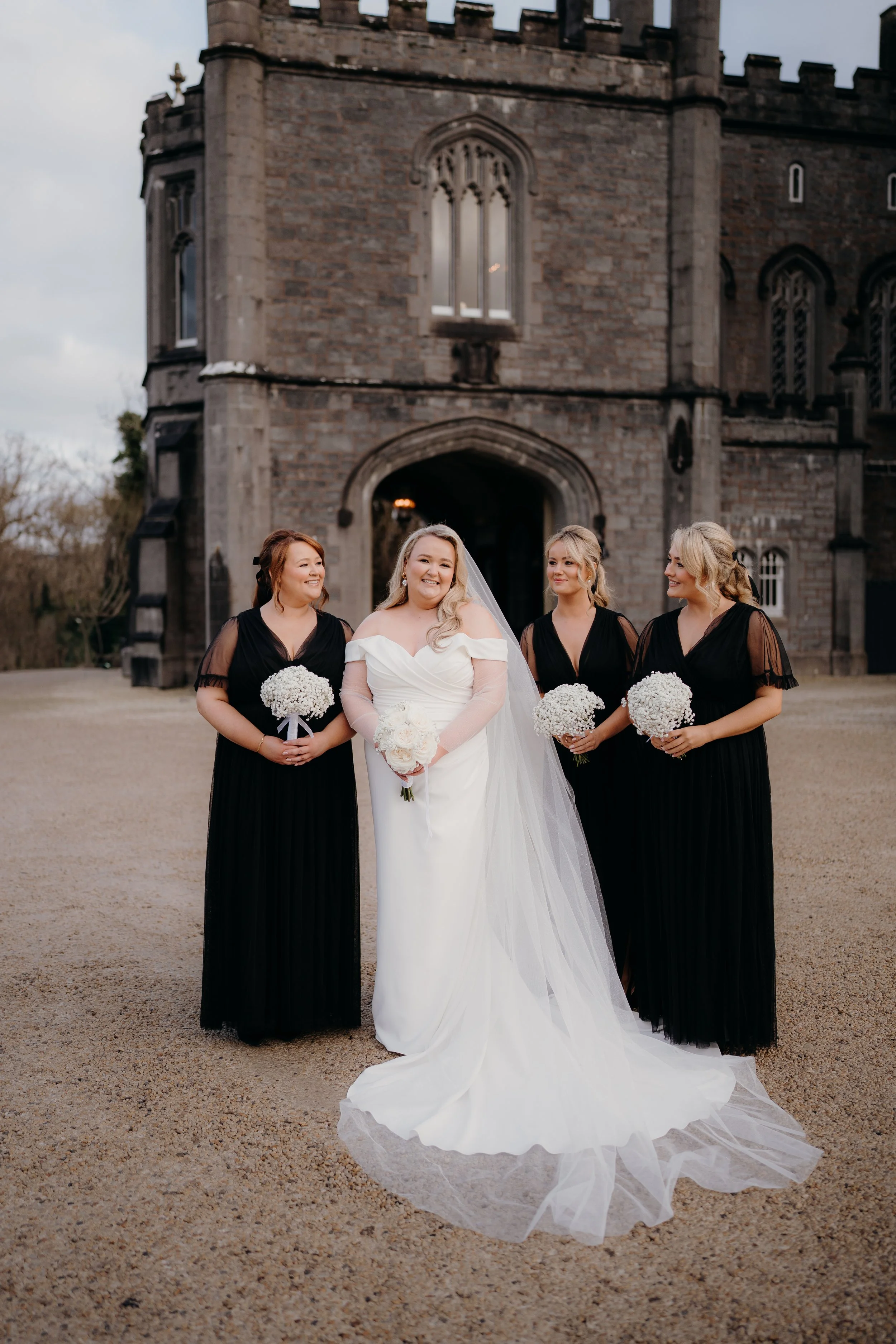 A bride in a white wedding dress holding a bouquet, standing with three bridesmaids in black dresses holding white flower bouquets, in front of a stone castle.