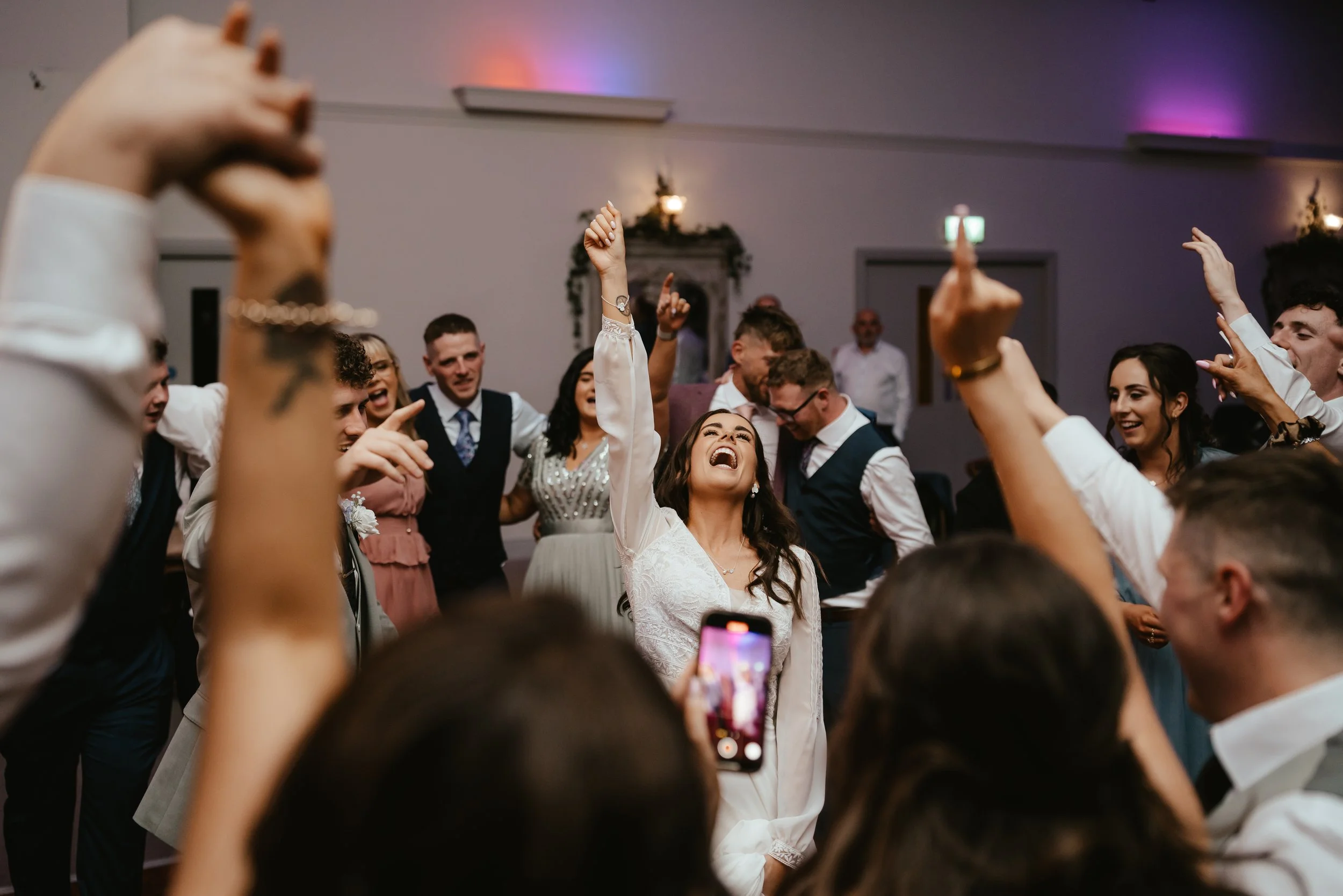 Group of people dancing and celebrating at a wedding reception, with a woman in a white dress smiling and raising her arm in the center.
