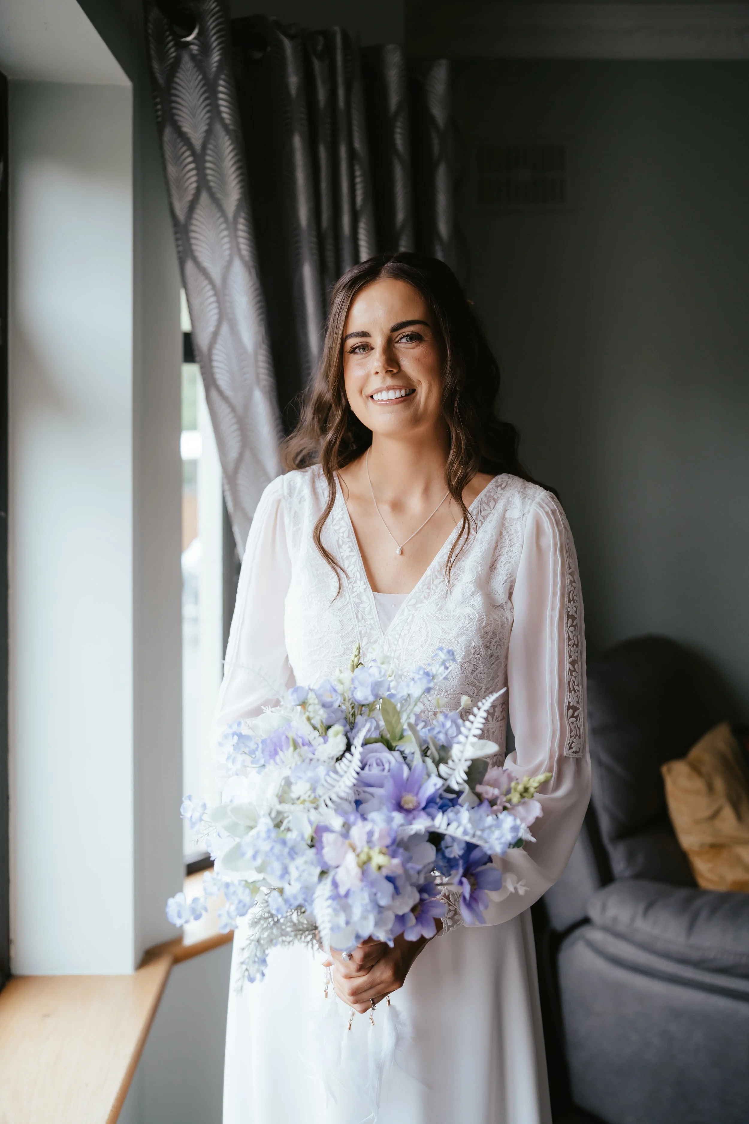 A woman in a white dress holding a bouquet of purple and white flowers, standing near a window with patterned curtains, smiling.