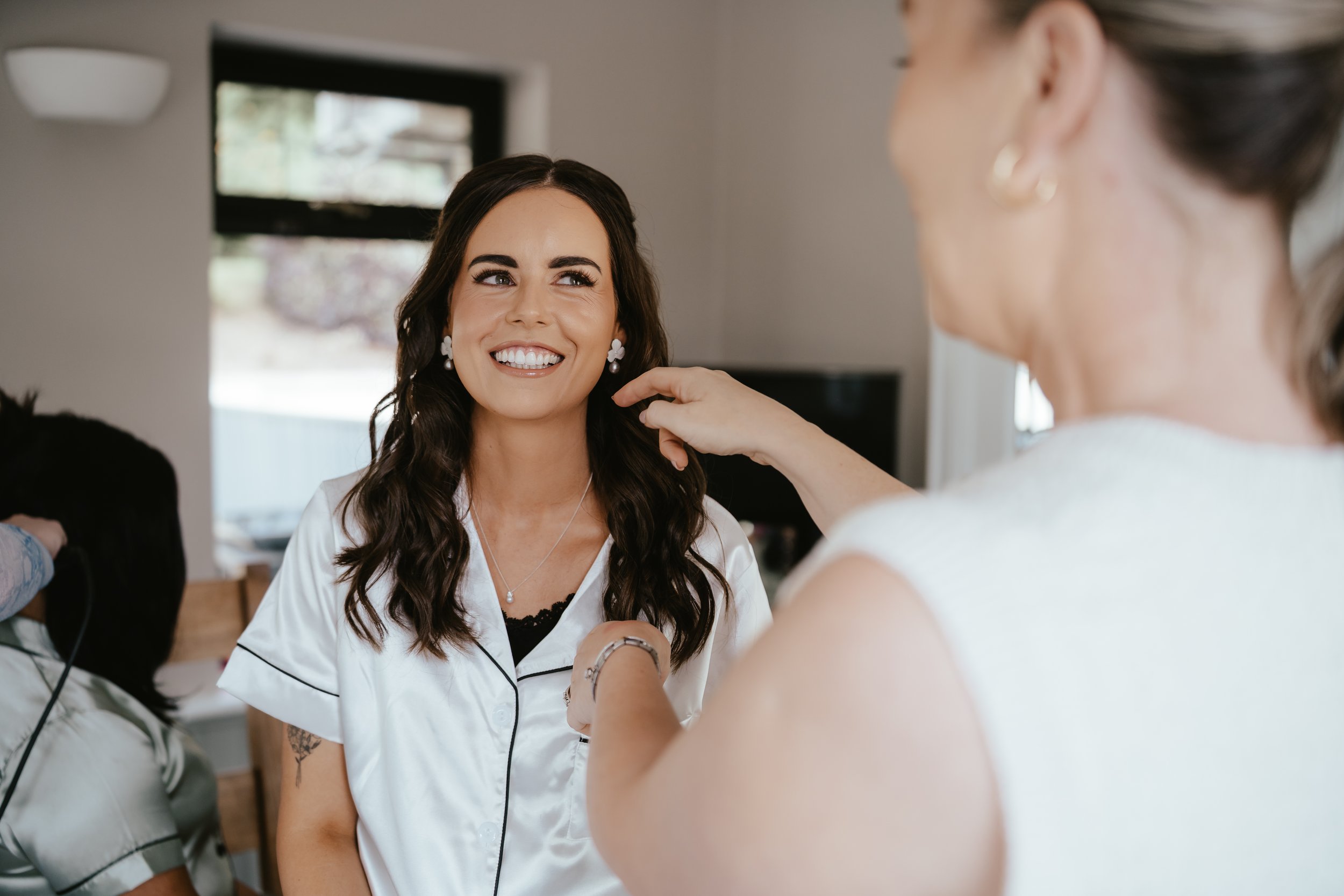 A woman with dark wavy hair during her makeup or hair styling, smiling while a makeup artist or stylist touches her face, in a well-lit room with a window in the background.