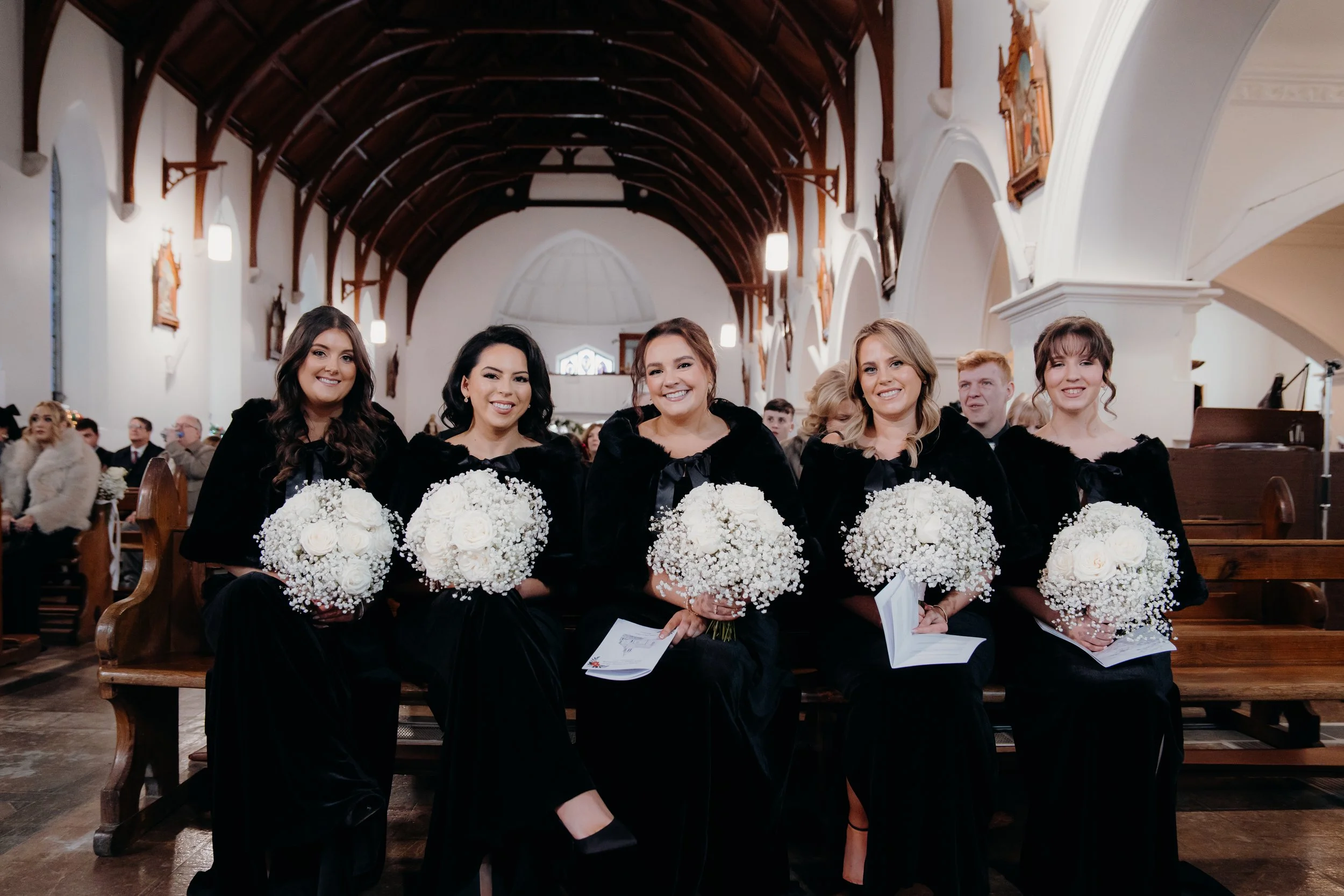 Women sitting in church on a wedding day, holding bouquets of white flowers, dressed in black dresses, inside a church with wooden beams and white walls.