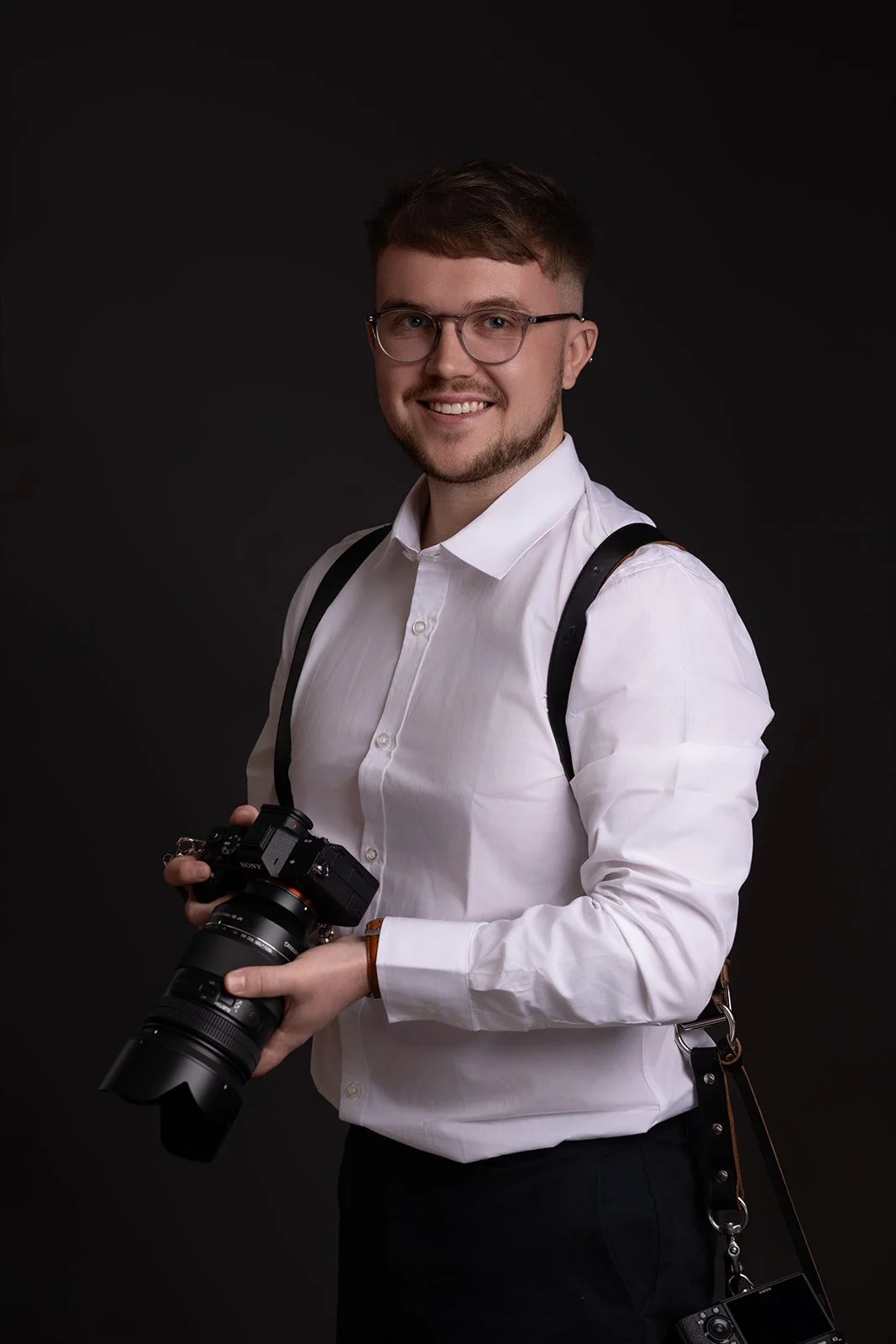 A young man with glasses, a beard, and a short haircut, wearing a white dress shirt and black pants, is smiling and holding a professional camera with a large lens while standing against a dark background.