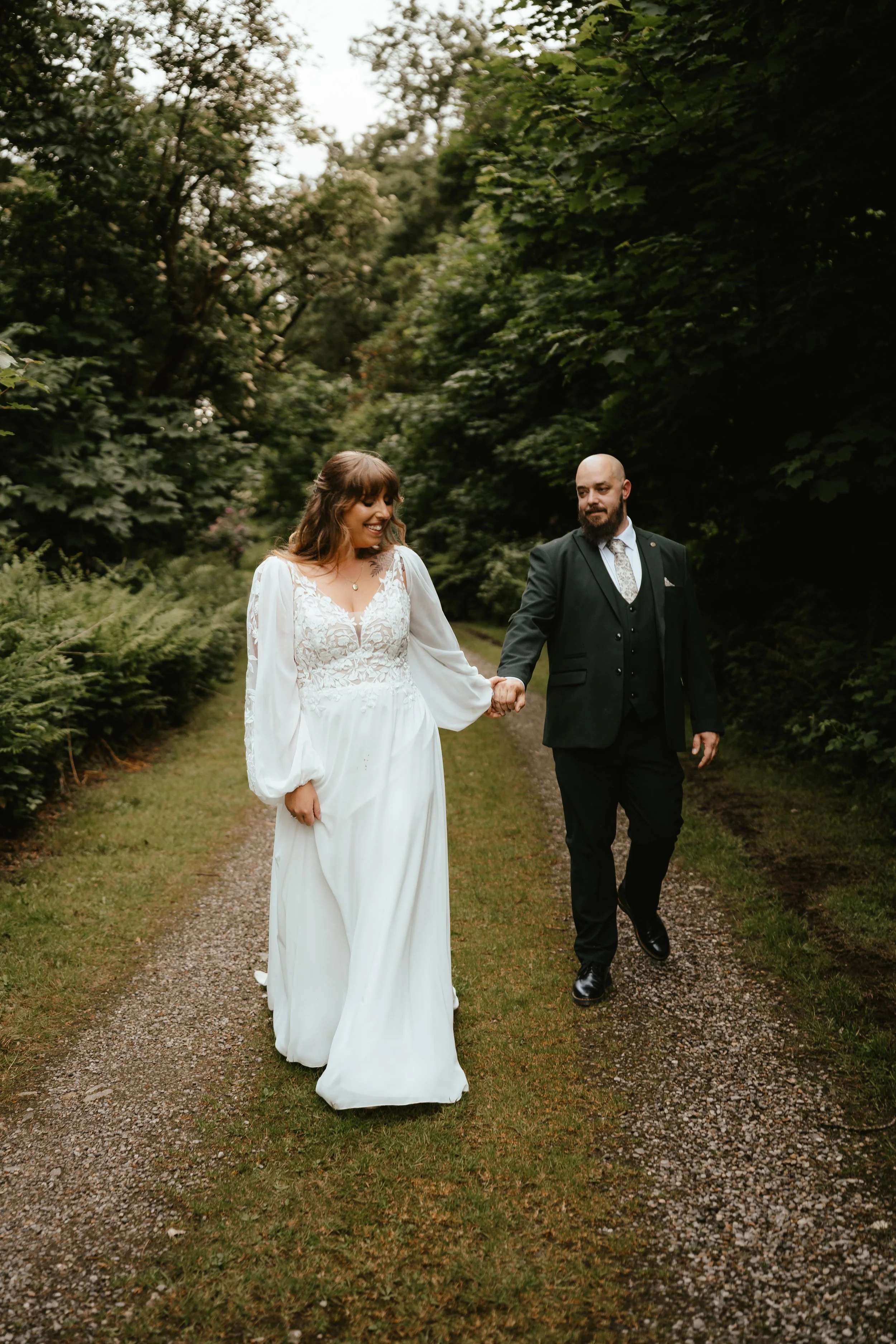 A newlywed couple walking hand in hand on a nature trail surrounded by lush green trees. The bride is in a white wedding dress with lace details, and the groom is in a dark suit with a patterned tie. They appear happy and relaxed.