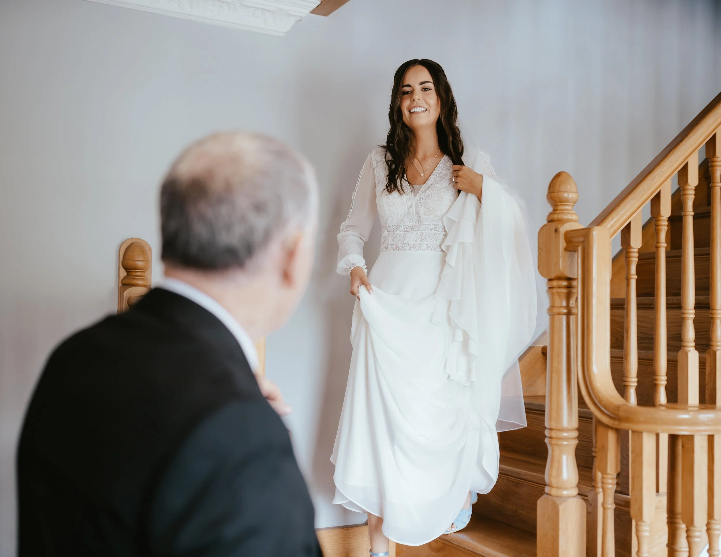 A woman in a white wedding dress smiling as she descends a wooden staircase, with an older man in a black suit watching from below.