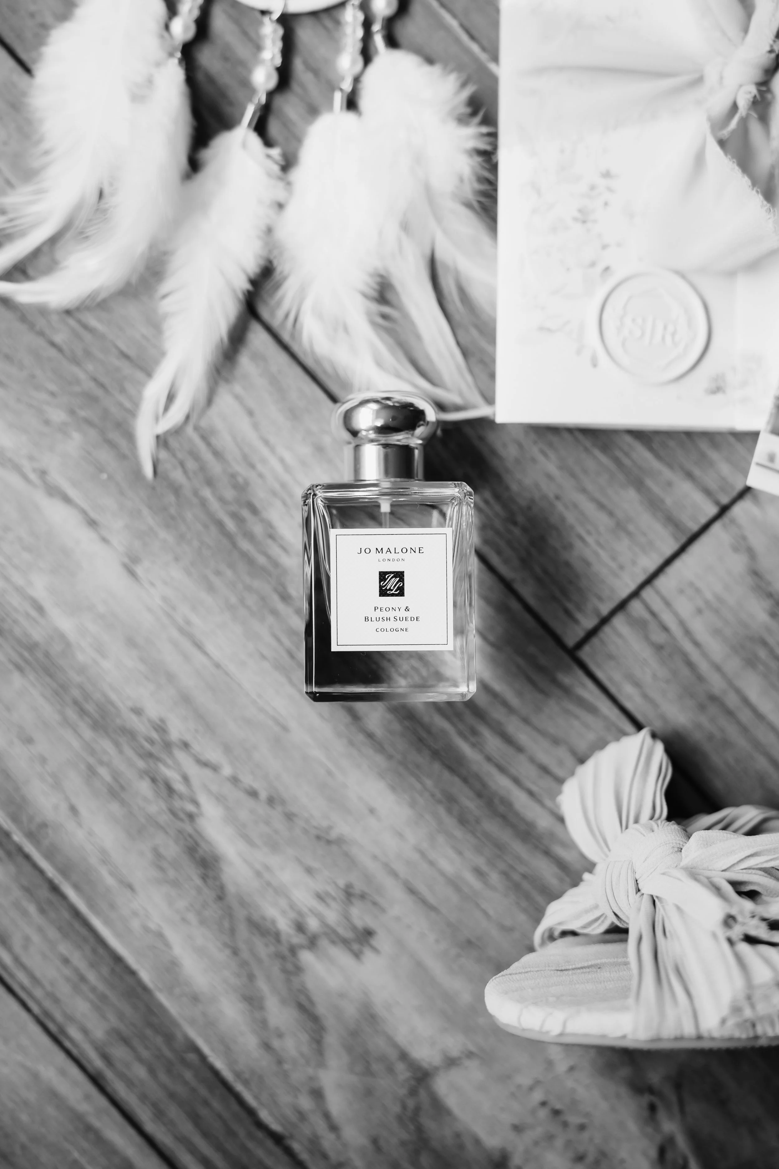 Black and white photo of a Jo Malone perfume bottle labeled 'Peony & Blush Suede Cologne' on a wooden surface, with feathers and paper around it.
