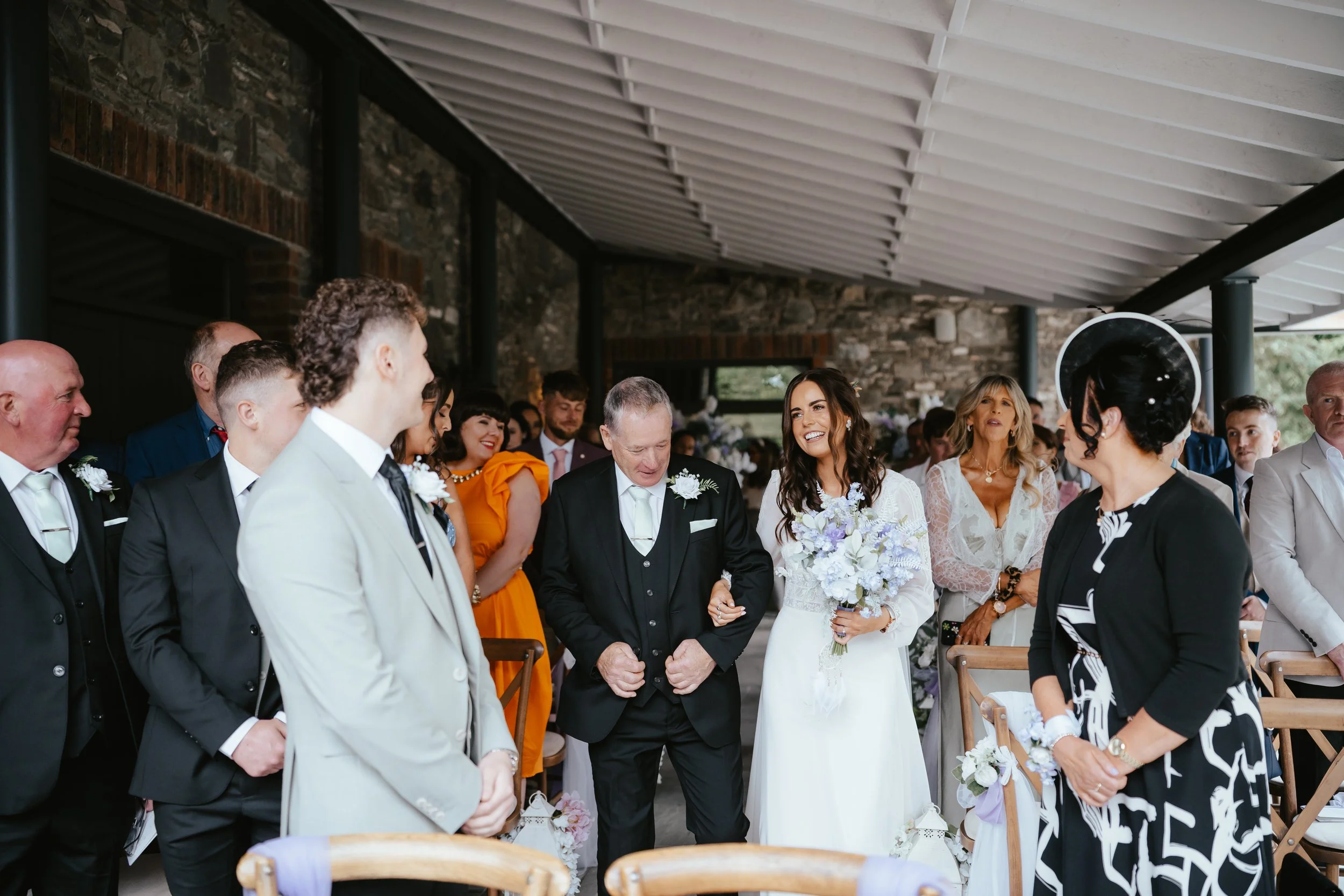 Bride walking down the aisle with her father, smiling, holding a bouquet of white and purple flowers, surrounded by wedding guests in an indoor ceremony with a brick wall and gray ceiling.