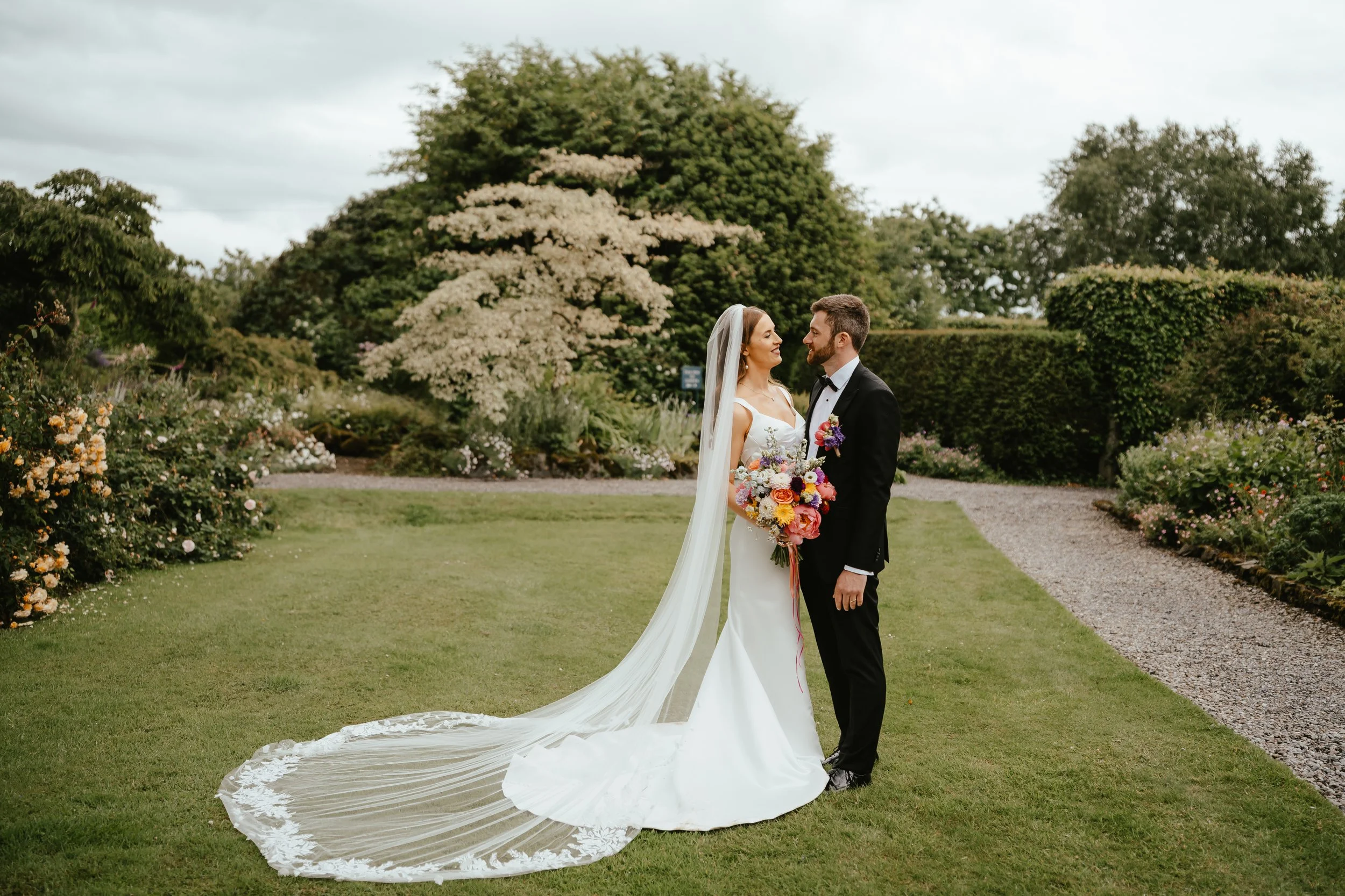 Bride and groom standing close together on a grassy path in a garden, smiling at each other. The bride wears a white wedding gown with a long train and veil, holding a colorful bouquet. The groom is dressed in a black tuxedo with a bow tie, also hold