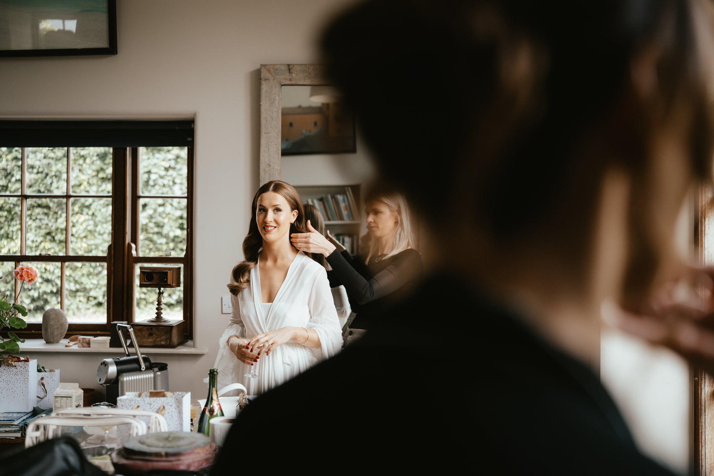 A woman in a white dress getting her hair styled by a hairstylist in a bright room with a window and shelves.