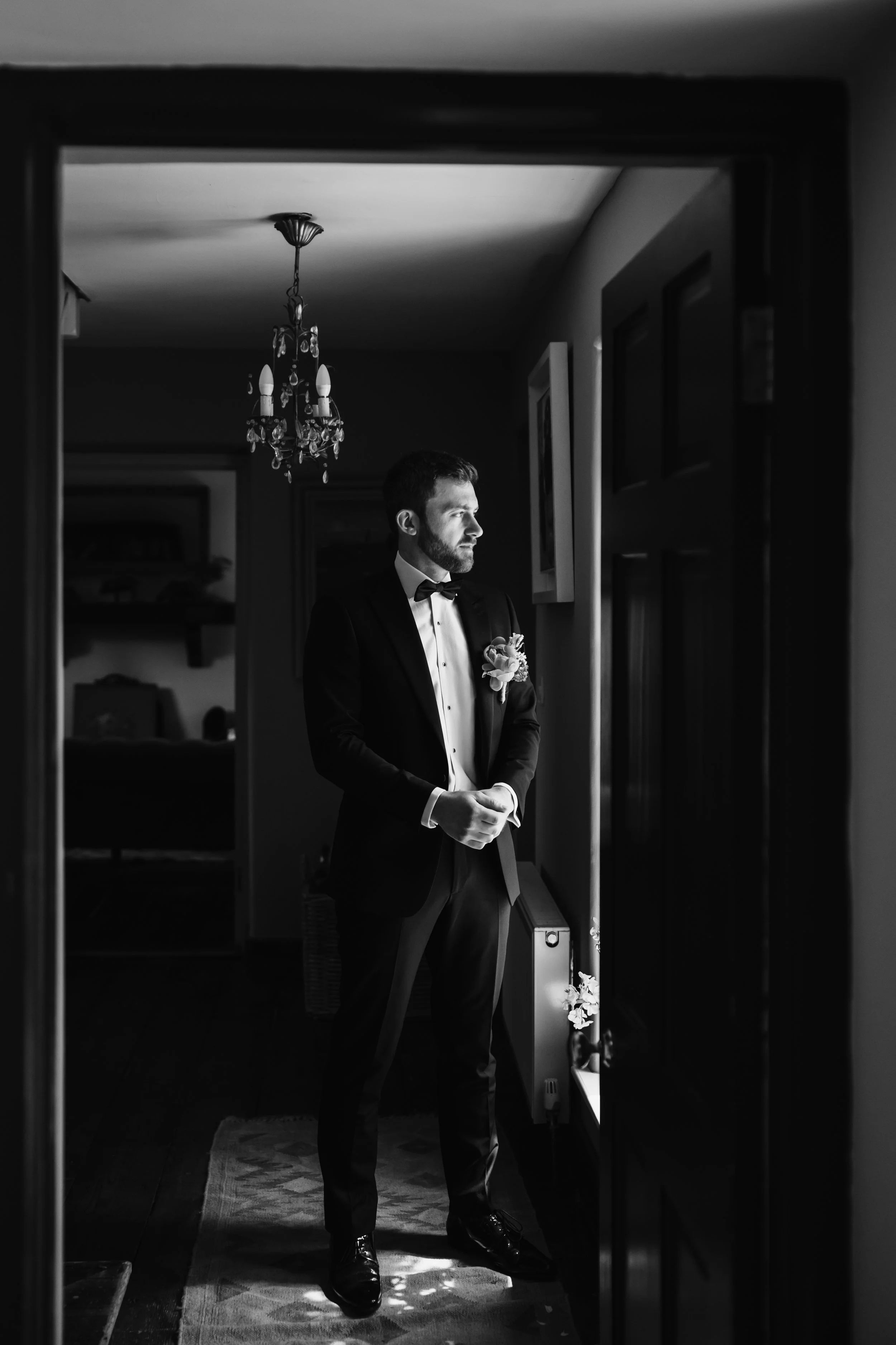 A man in formal tuxedo with a bow tie and boutonniere, standing and looking out a window in a dimly lit room, with a chandelier overhead and artwork on the wall.