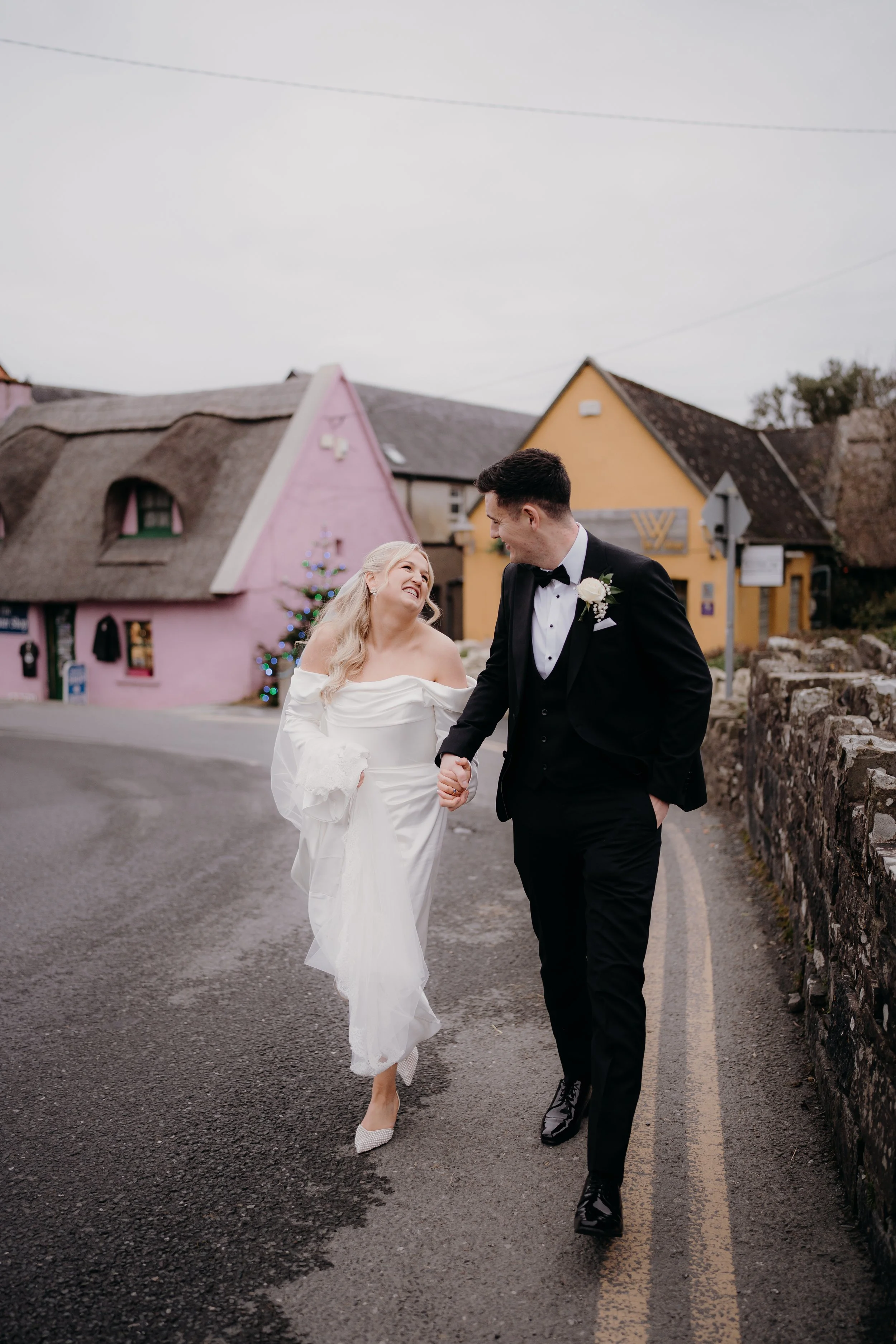 A newlywed couple walking hand in hand outdoors on a cloudy day, with colorful houses in the background. The bride is wearing a white off-the-shoulder wedding gown and the groom is dressed in a black tuxedo with a bow tie.