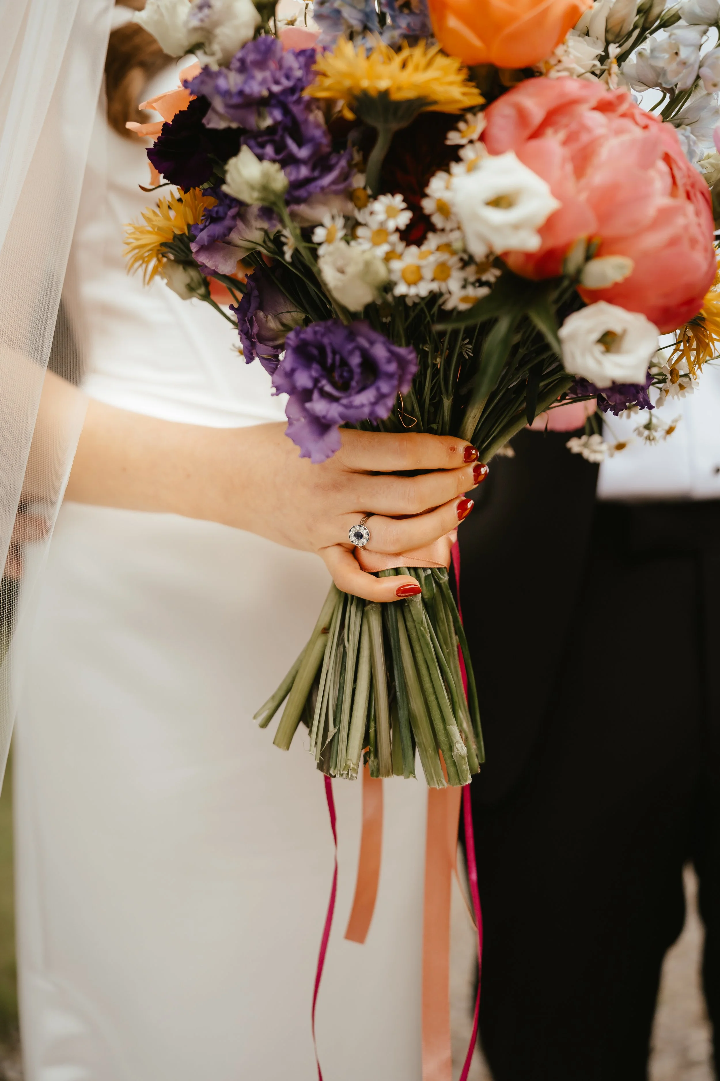 A woman in a white dress holding a colorful bouquet of flowers including purple, white, yellow, and pink blossoms, with a wedding ring on her finger.
