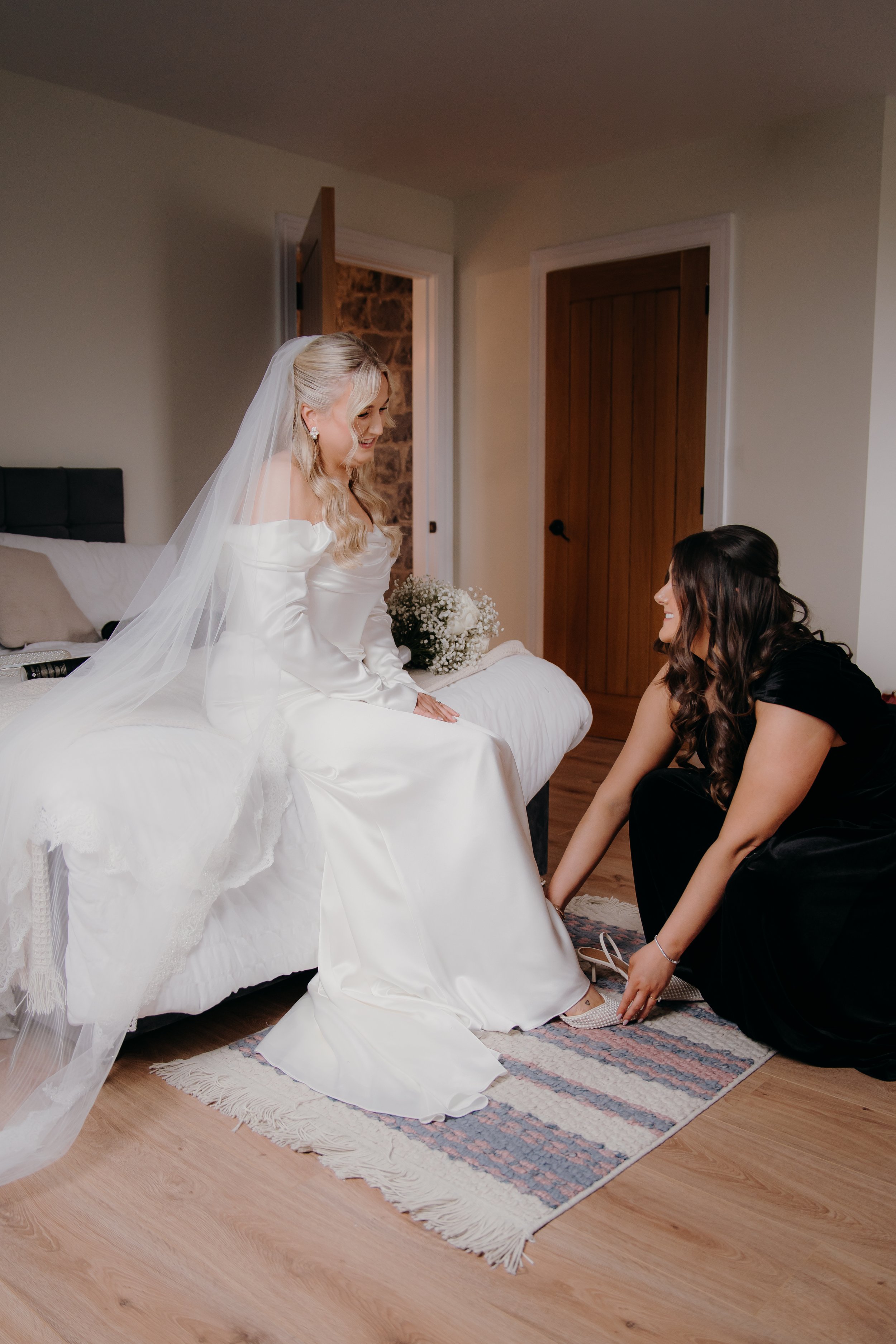 Bridal bride sitting on bed, smiling at her bridesmaid who is helping her with her shoe in a bedroom.