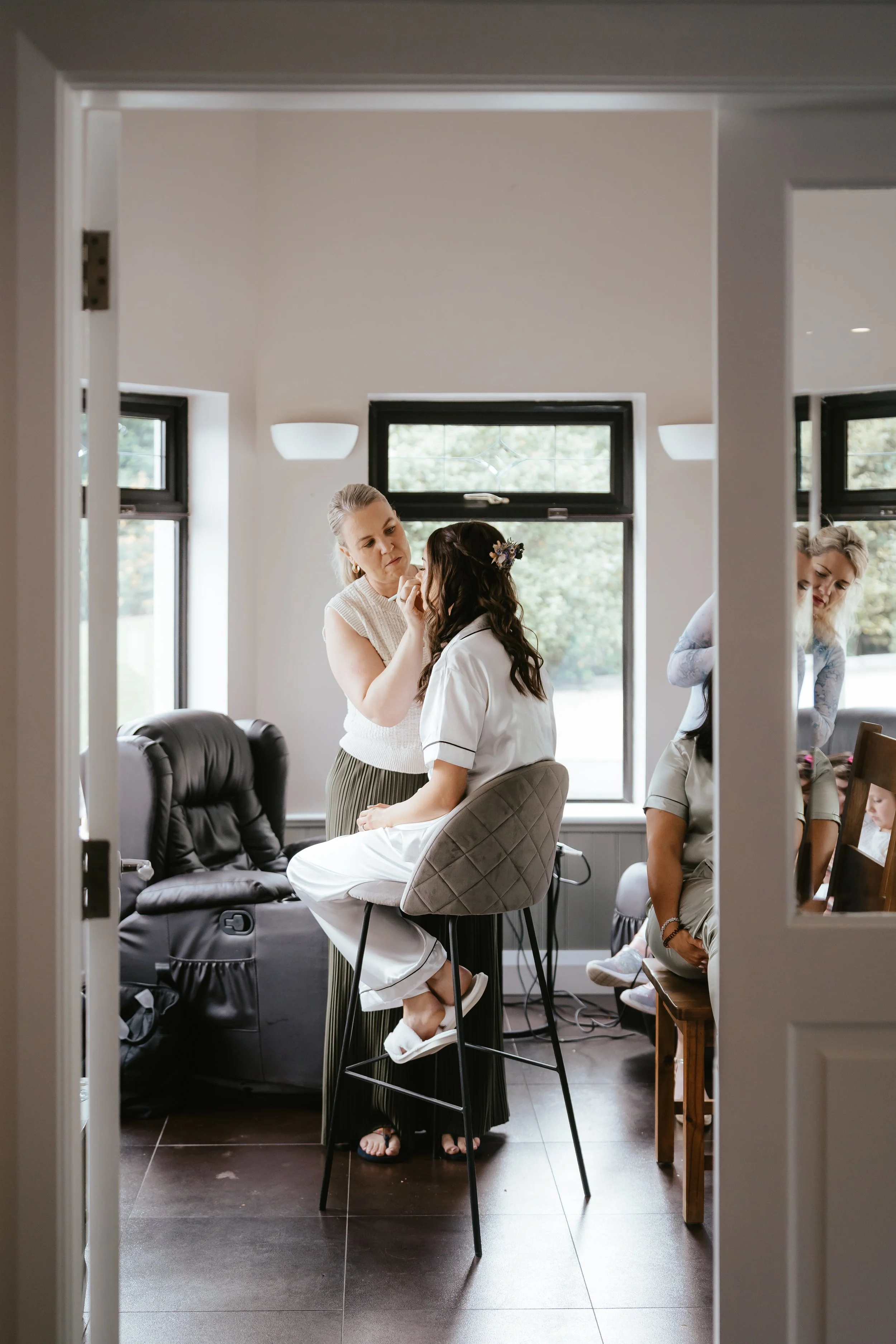 A woman sitting on a high chair while another woman applies makeup or skincare to her face in a room with large windows and natural light.