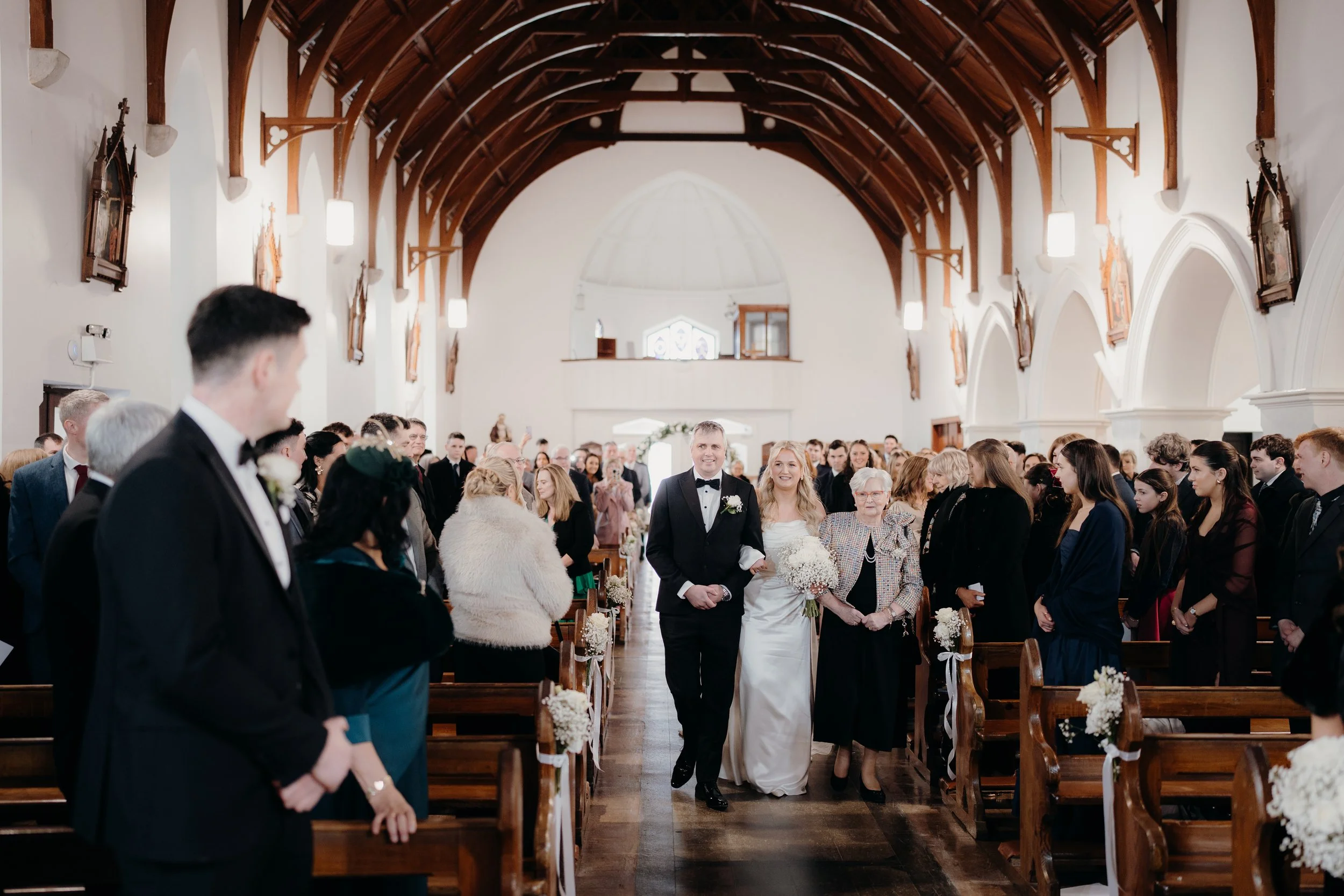 A bride and groom walking down the aisle inside a church, surrounded by guests. The bride is holding a bouquet and smiling, while the groom is dressed in a tuxedo. The church has wooden beams and religious portraits on the walls.
