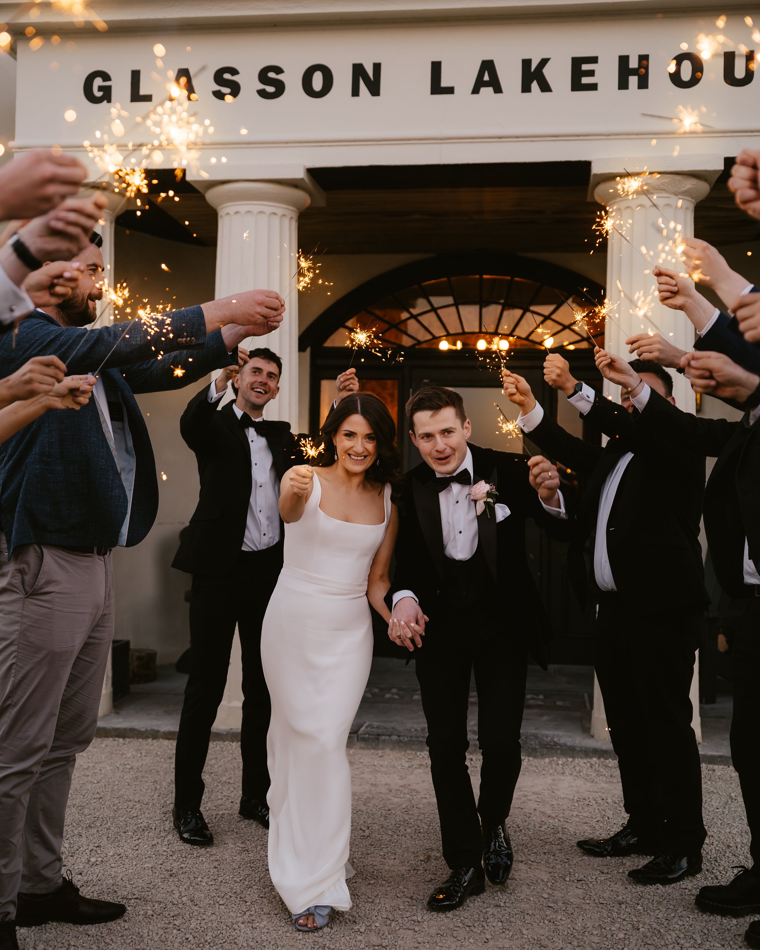 A newly married couple holding hands and smiling as they walk through a crowd of people holding sparklers outside Glisson Lakehouse.