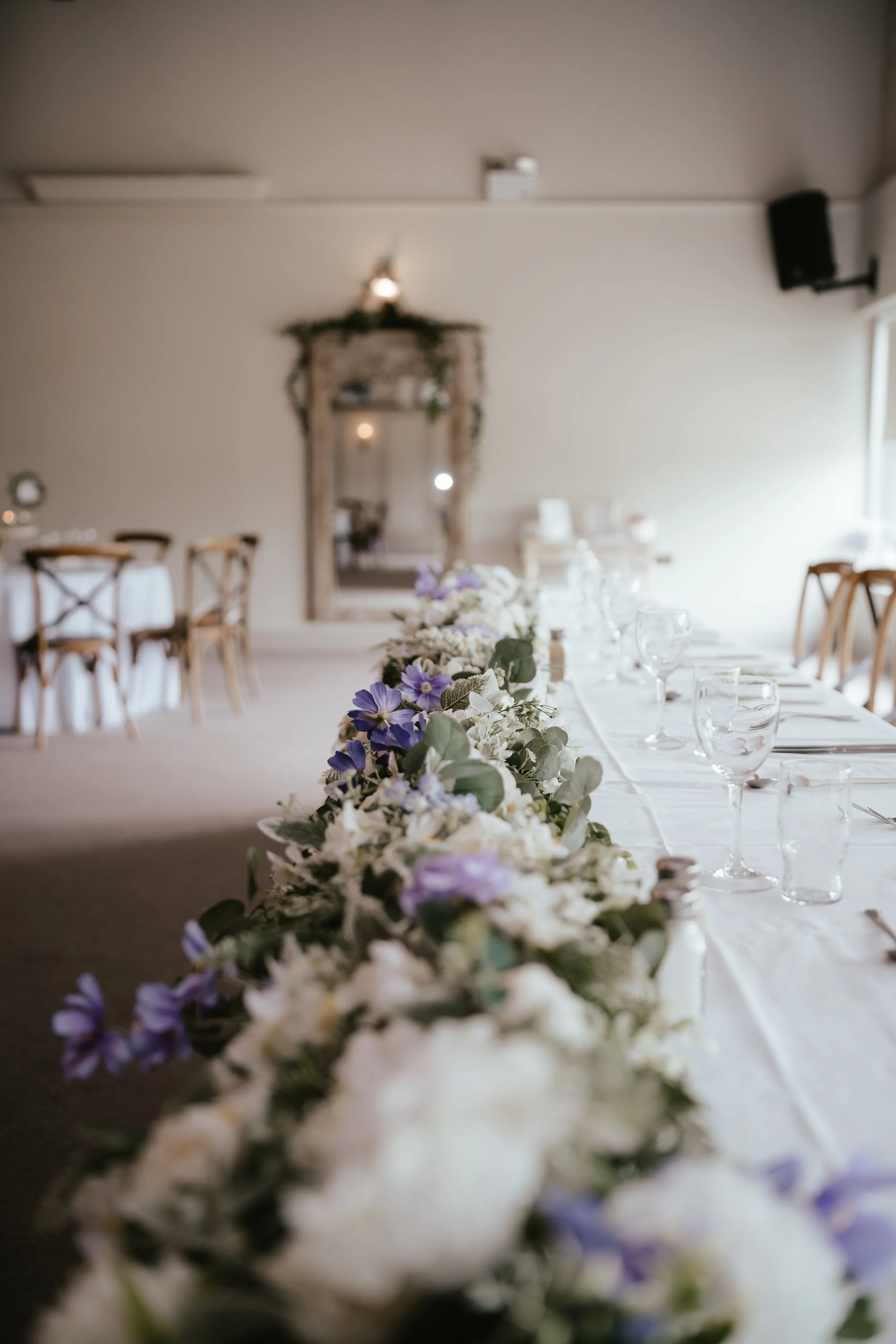 A decorated event table with a floral garland, set with glassware and cutlery, in a softly lit room with a mirror and chairs in the background.