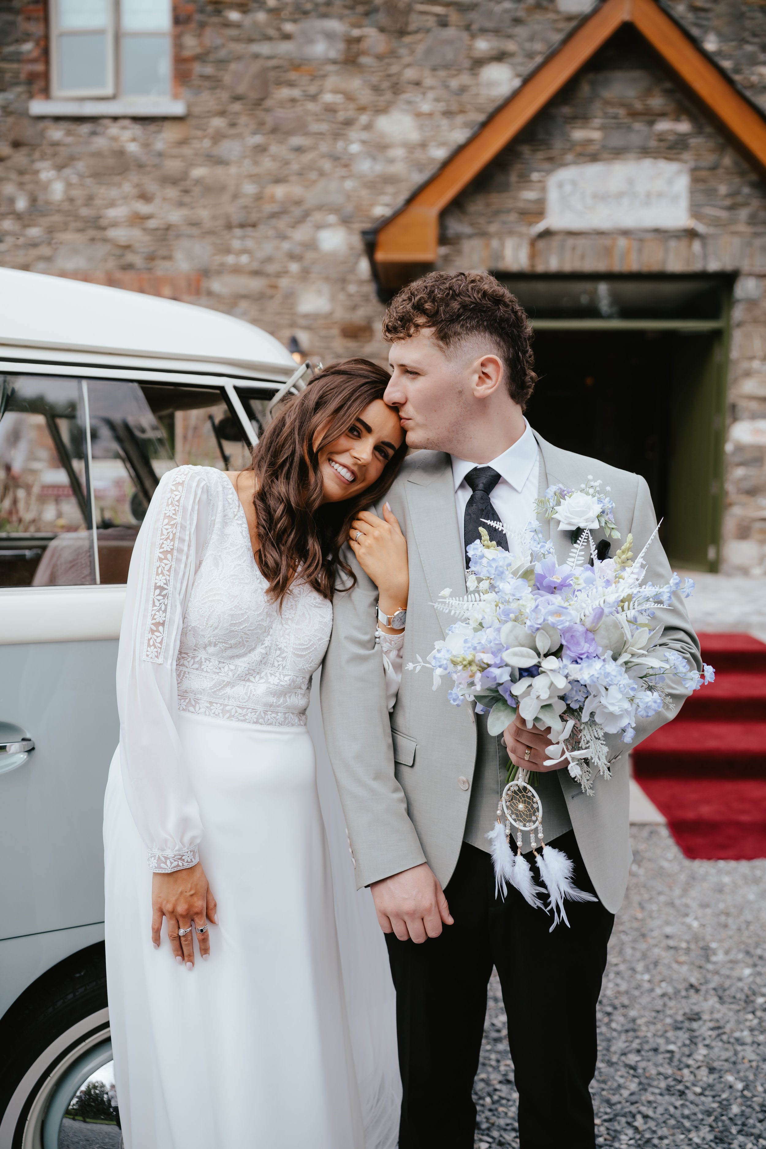 A bride and groom stand close together outside a stone building, with the groom holding a bouquet of white and purple flowers. The bride is leaning her head on the groom's forehead, smiling happily. The groom appears to kiss the bride's forehead.