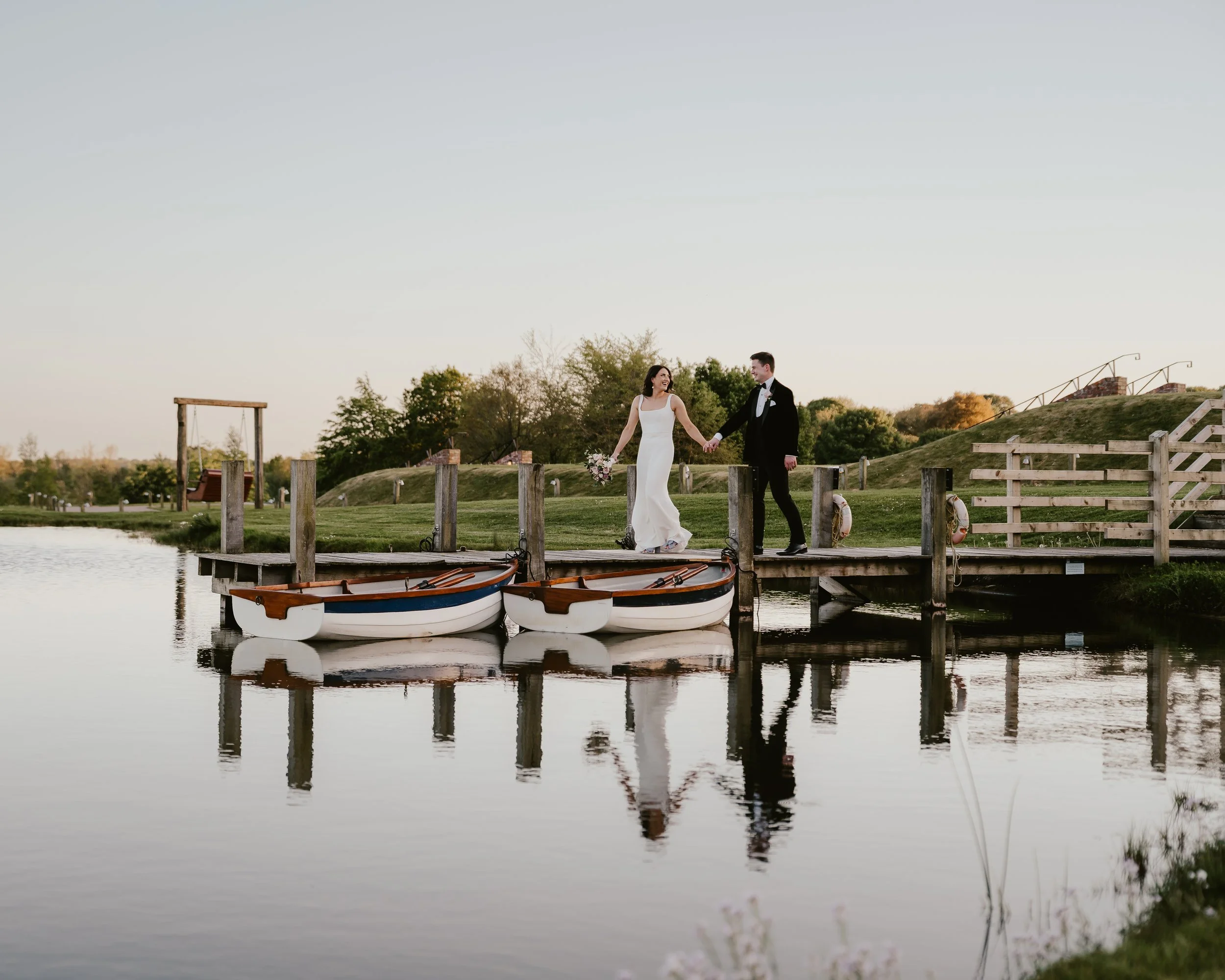 Bride and groom holding hands on a wooden dock over water, with small boats reflected in the water, and trees and grassy hills in the background during sunset.