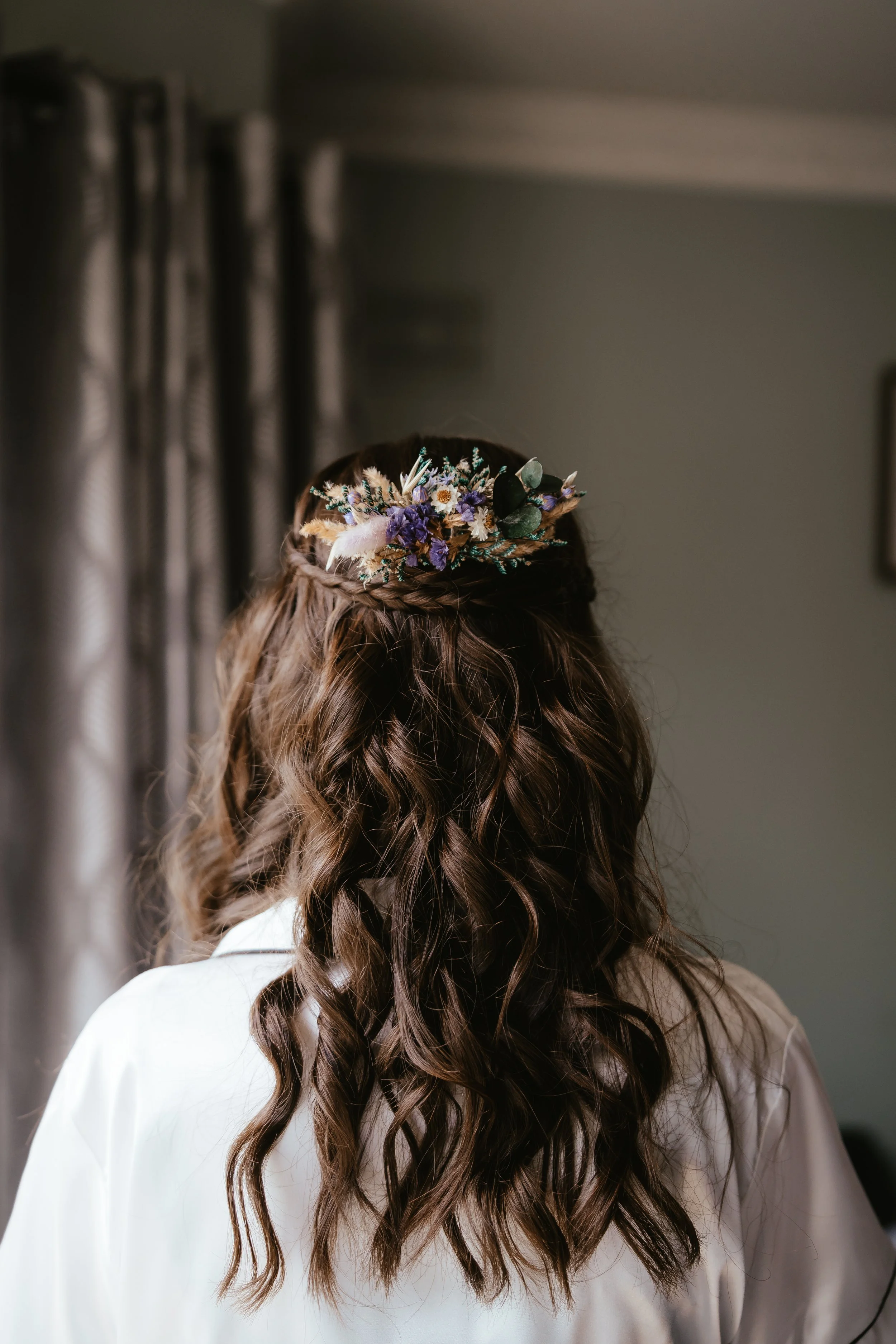 Back view of a woman with wavy brown hair wearing a hairpiece with purple, white, and green flowers and a white shirt, standing in a room with grey curtains.