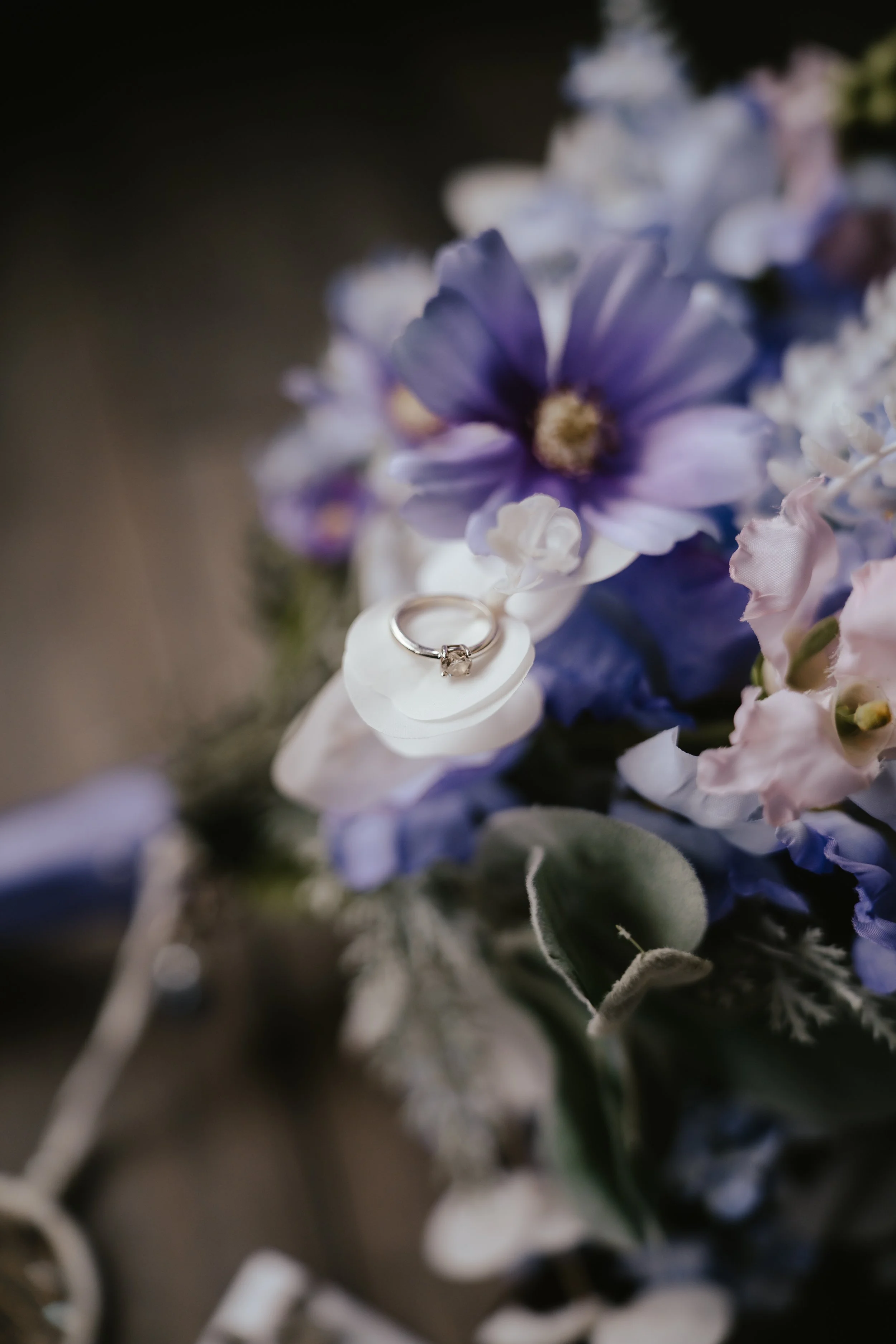 A silver engagement ring with a square-cut diamond rests on a white petal surrounded by purple, pink, and white flowers, with green leaves in the background.
