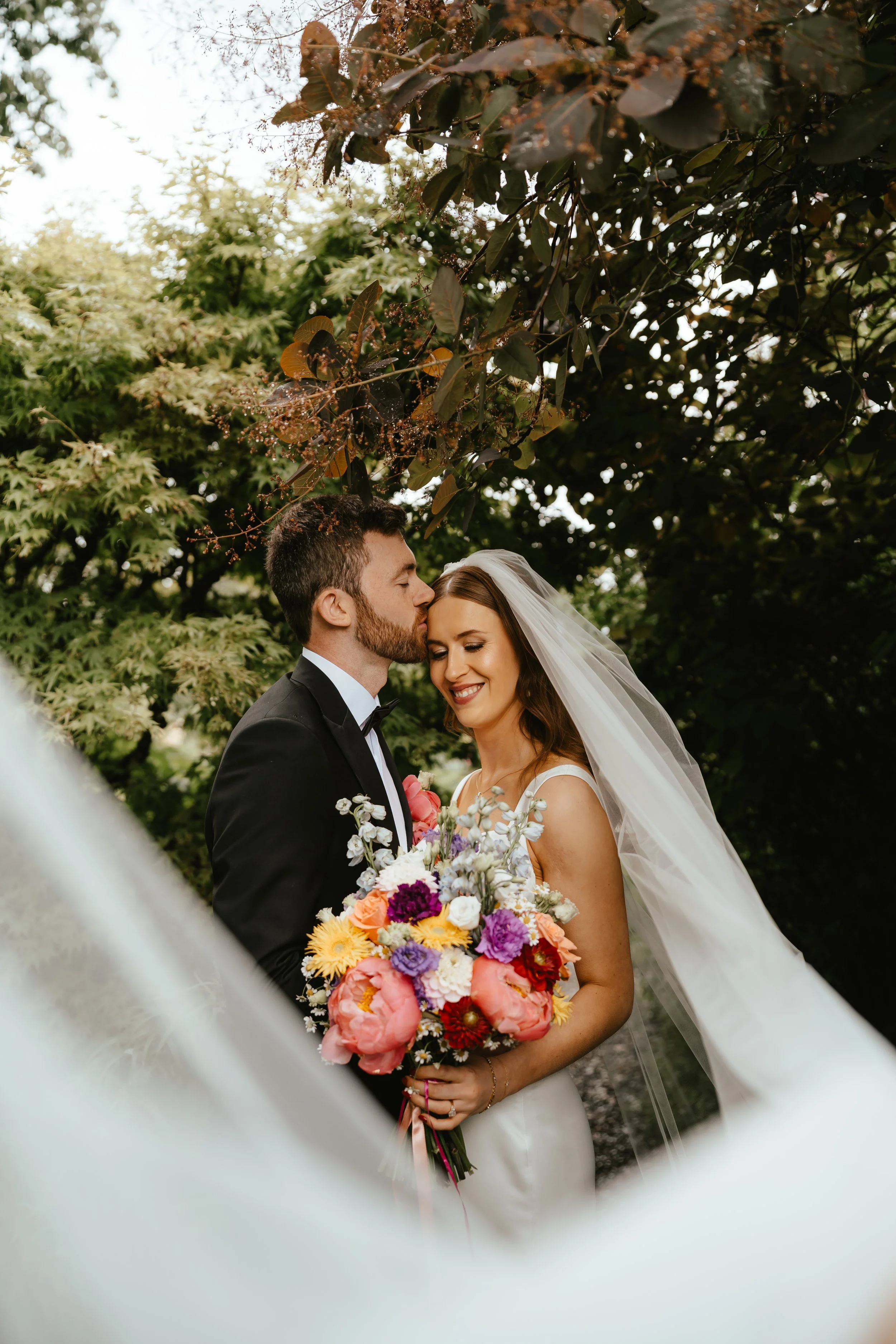 A bride and groom on their wedding day, surrounded by greenery. The groom is kissing the bride's forehead, and she is smiling while holding a colorful bouquet.