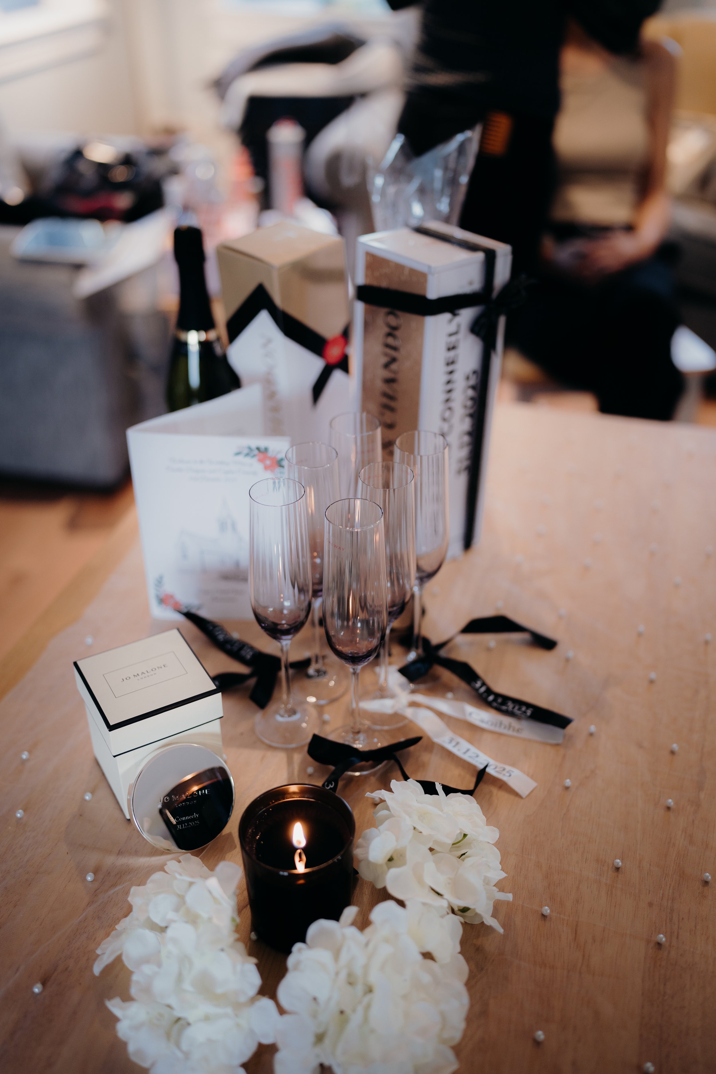 Table decorated with white flower arrangement, lit candle, empty champagne glasses, and gifts, in a cozy indoor setting.