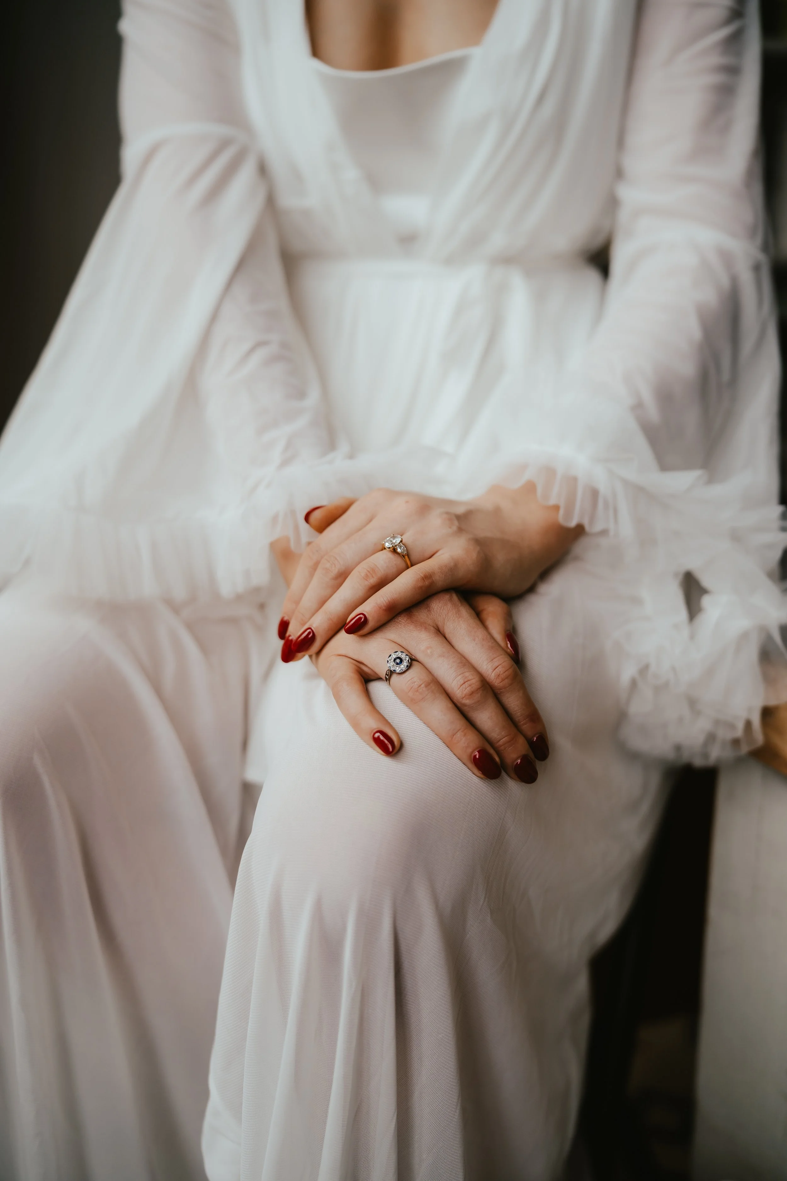 Close-up of a person's crossed hands resting on their lap, wearing a white dress with long sleeves and decorative ruffled cuffs. The person has red nail polish and is wearing rings, including one with a large gemstone and another with a smaller gemst
