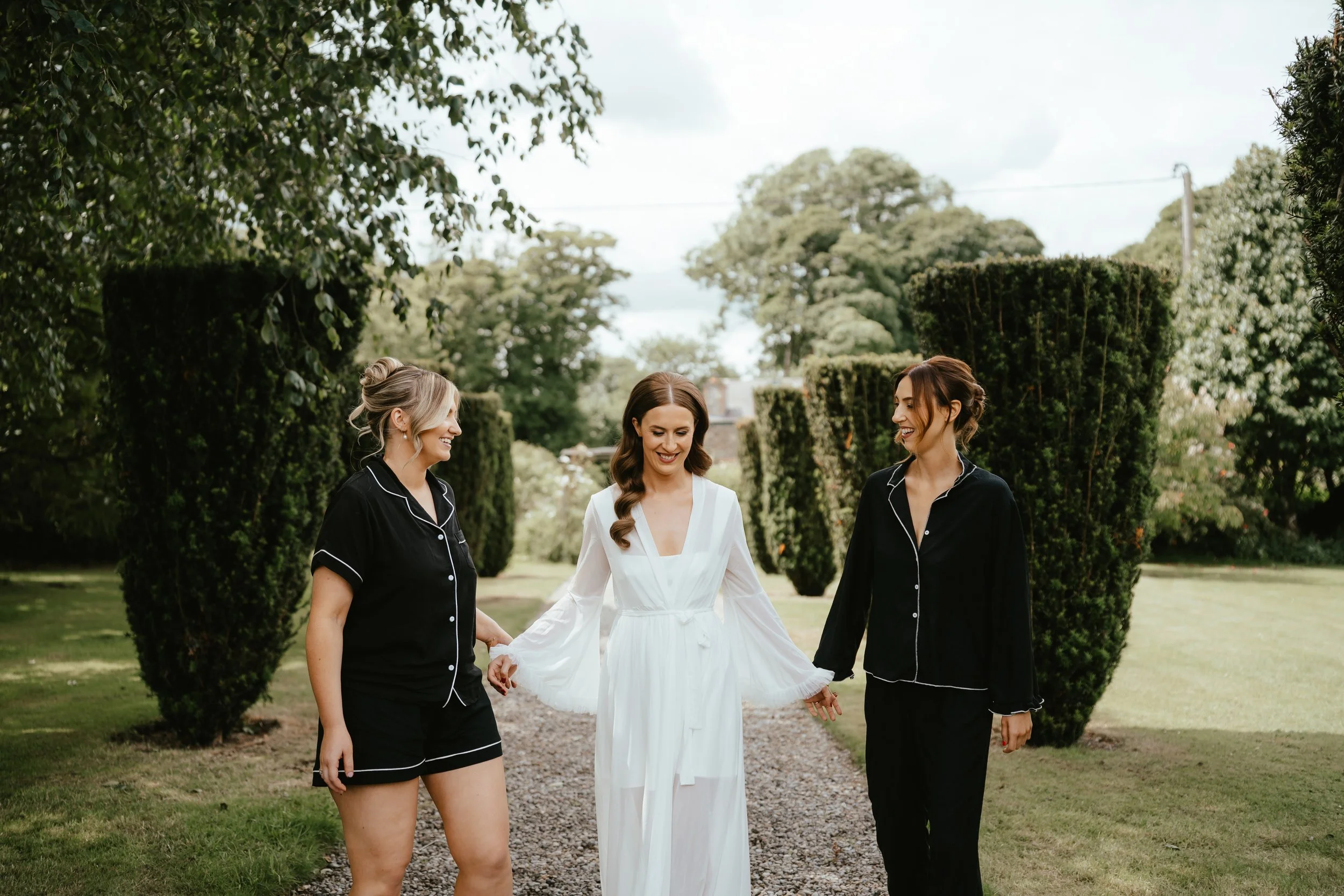 A bride in a white dress walking outdoors with two women in black pajamas, smiling and holding hands, in a garden with trimmed bushes and trees.