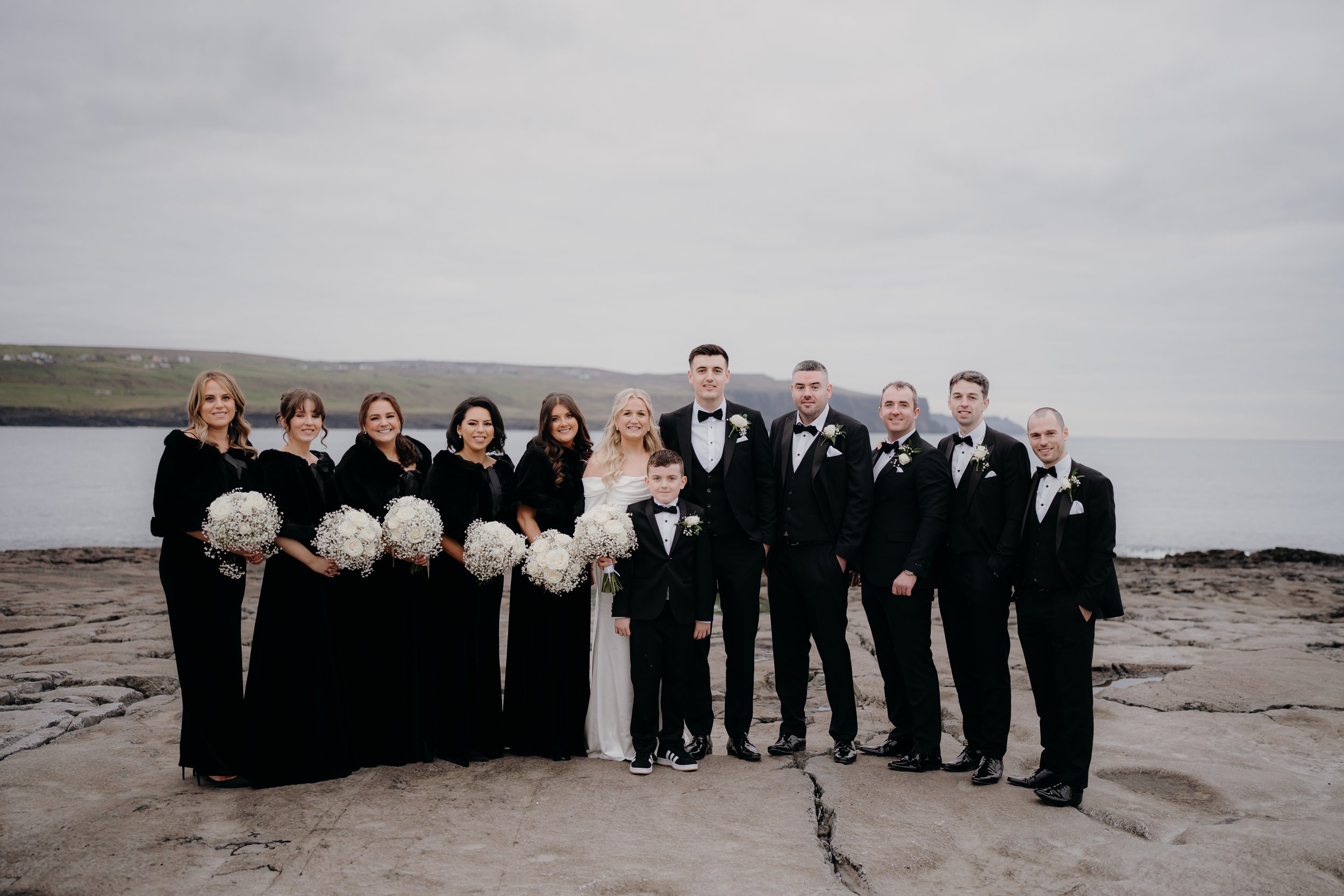 Wedding party standing on rocky shore by water, including bride, groom, bridesmaids, groomsmen, and children, all dressed in formal attire, with a cloudy sky in the background.