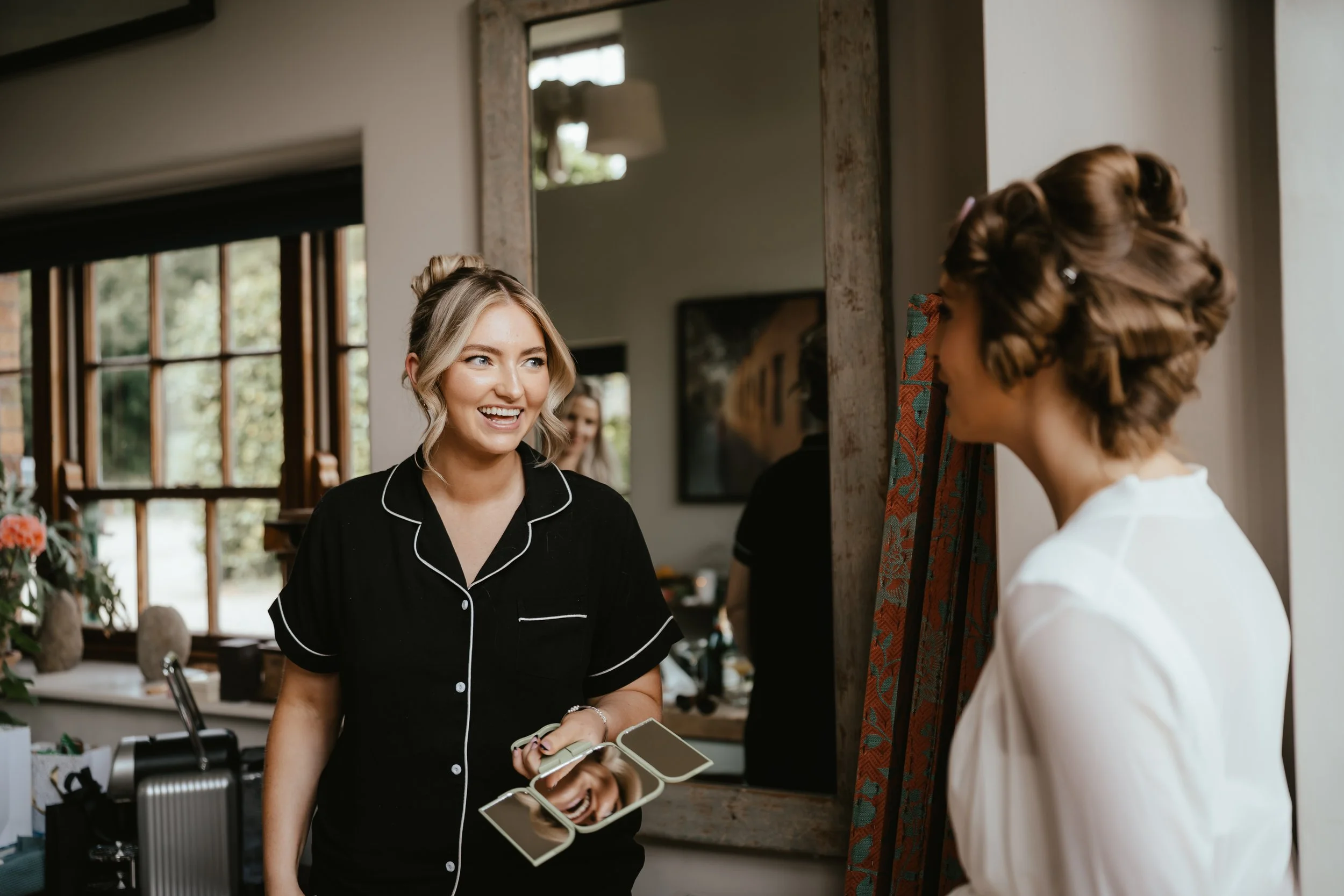Two women talking and smiling in a room with a large mirror, one holding a mirror and wearing black pajamas, the other wearing a white robe, with hairstyling in progress.