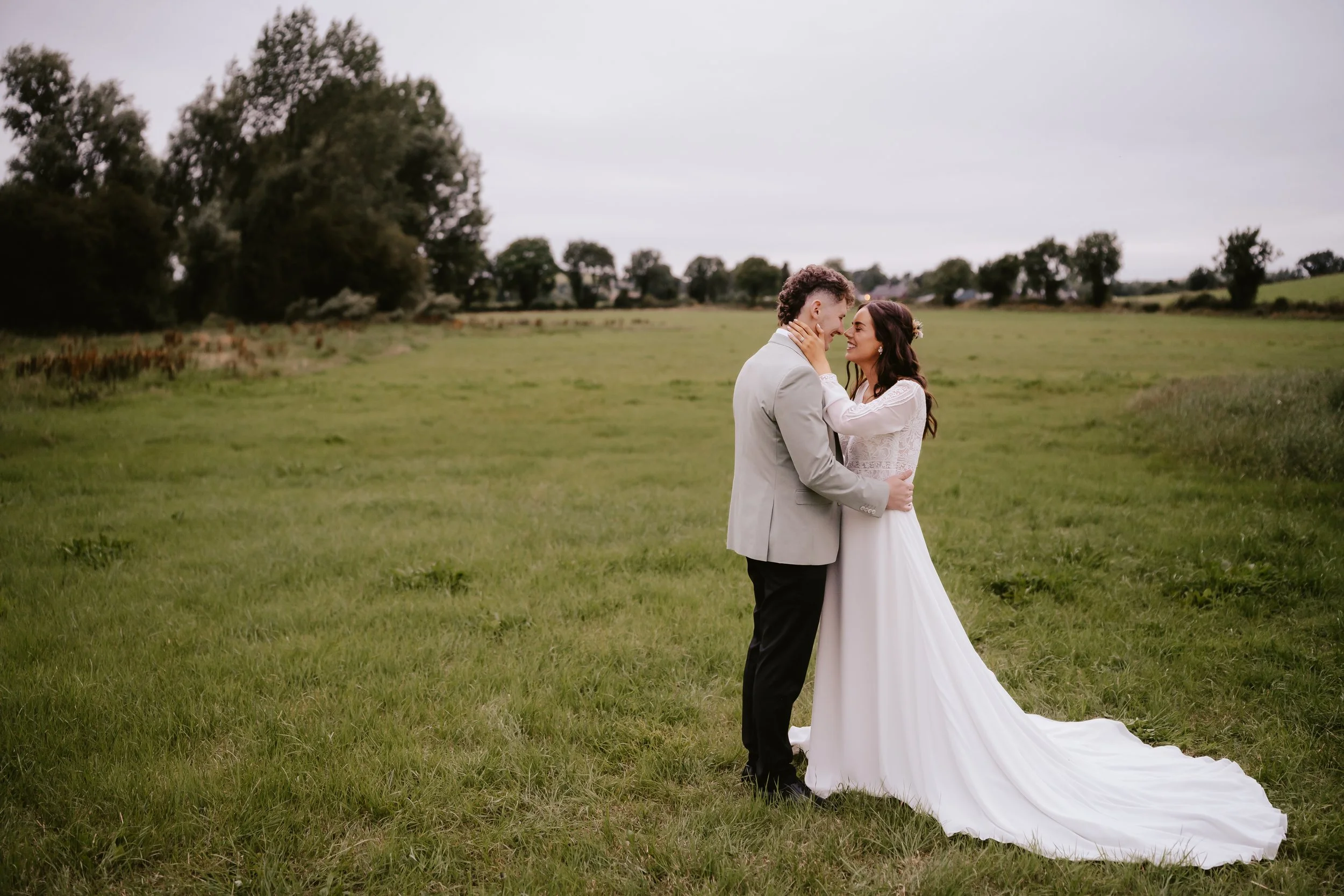 A bride and groom standing in a grassy field, gazing into each other's eyes, with trees and an overcast sky in the background.