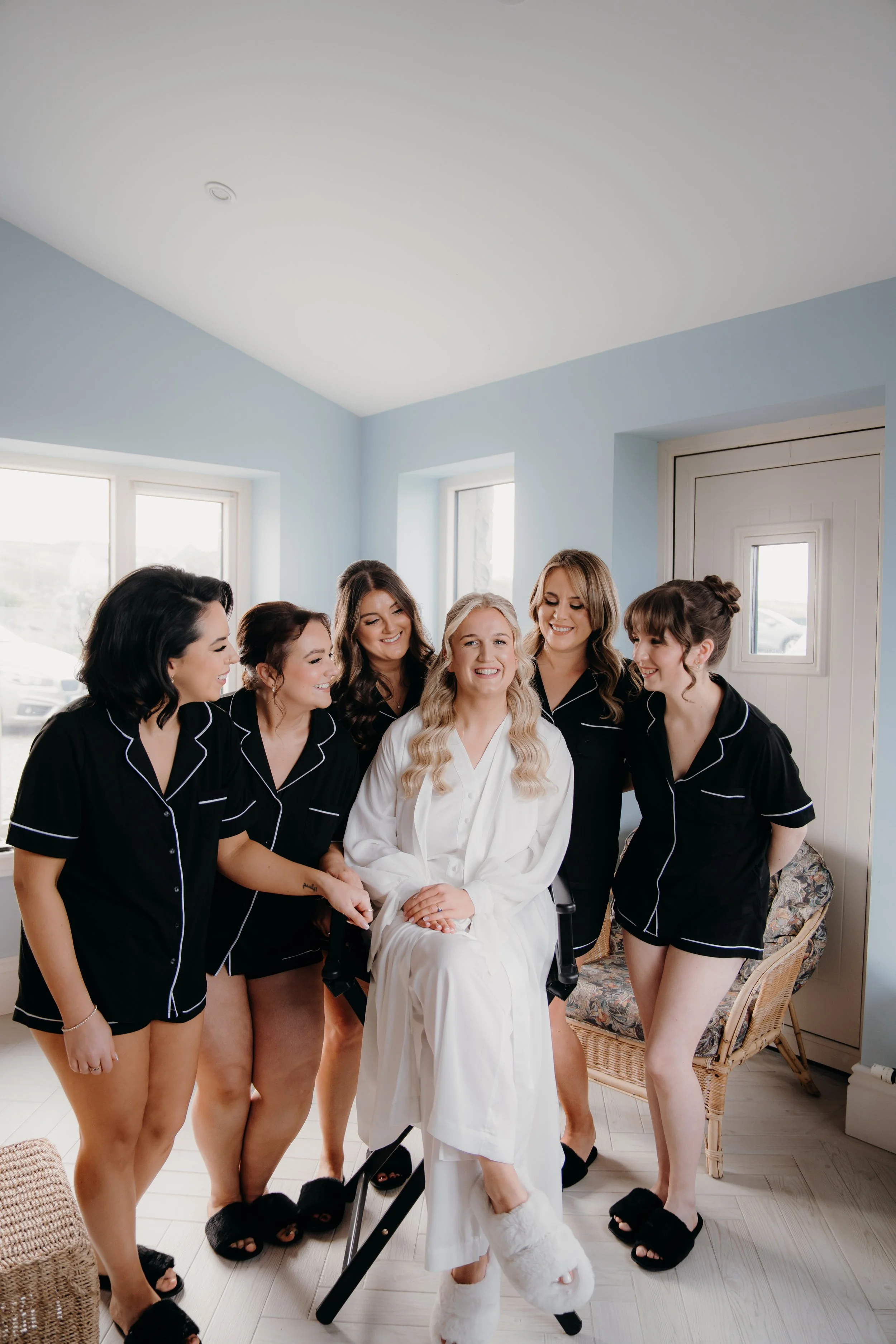 A bride in a white robe sitting on a chair surrounded by six bridesmaids in matching black pajamas, celebrating together in a well-lit room.