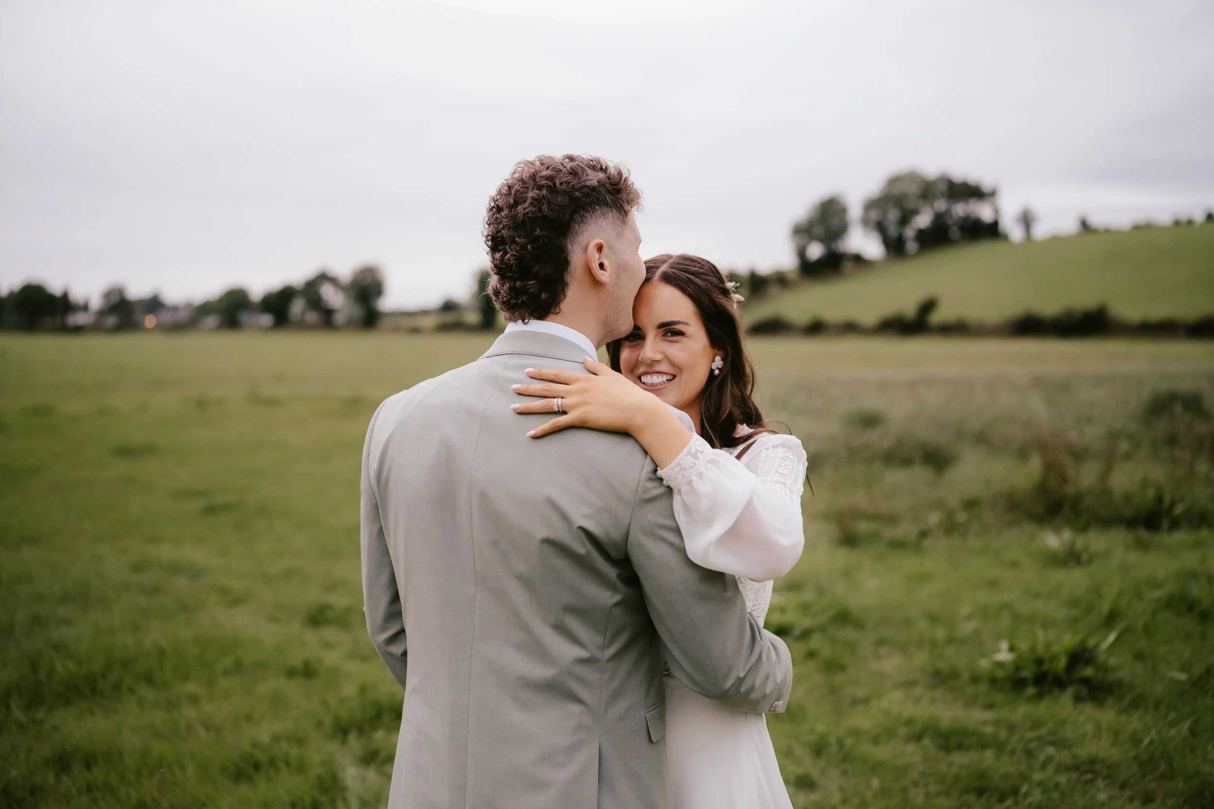 A newlywed couple embraces outdoors in a grassy field with rolling hills and trees in the background, smiling and sharing a moment of joy.
