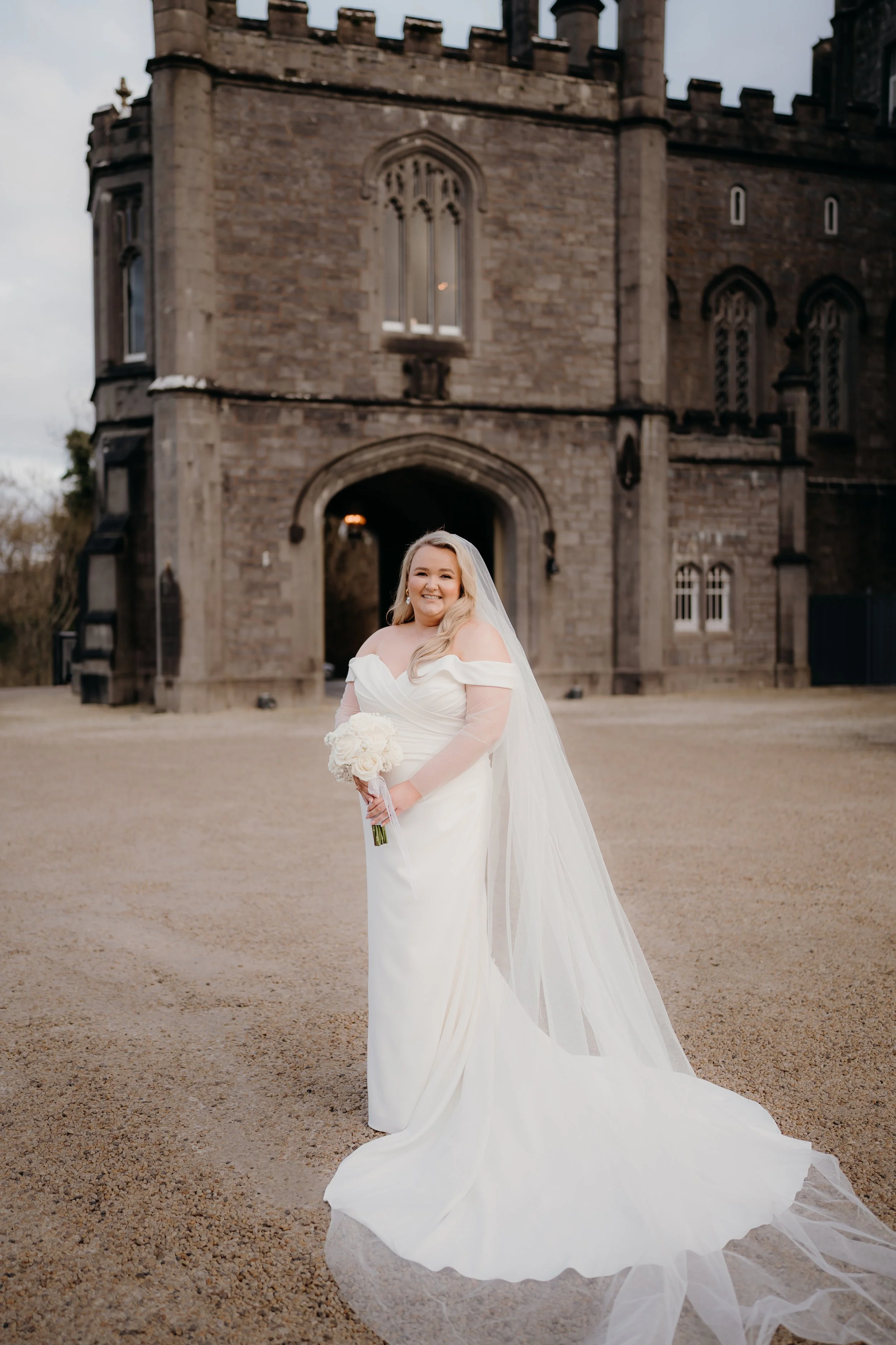 A bride in a white wedding dress and veil holding a bouquet of flowers, standing outdoors in front of a historic castle-like building with a stone facade and arched entrance.