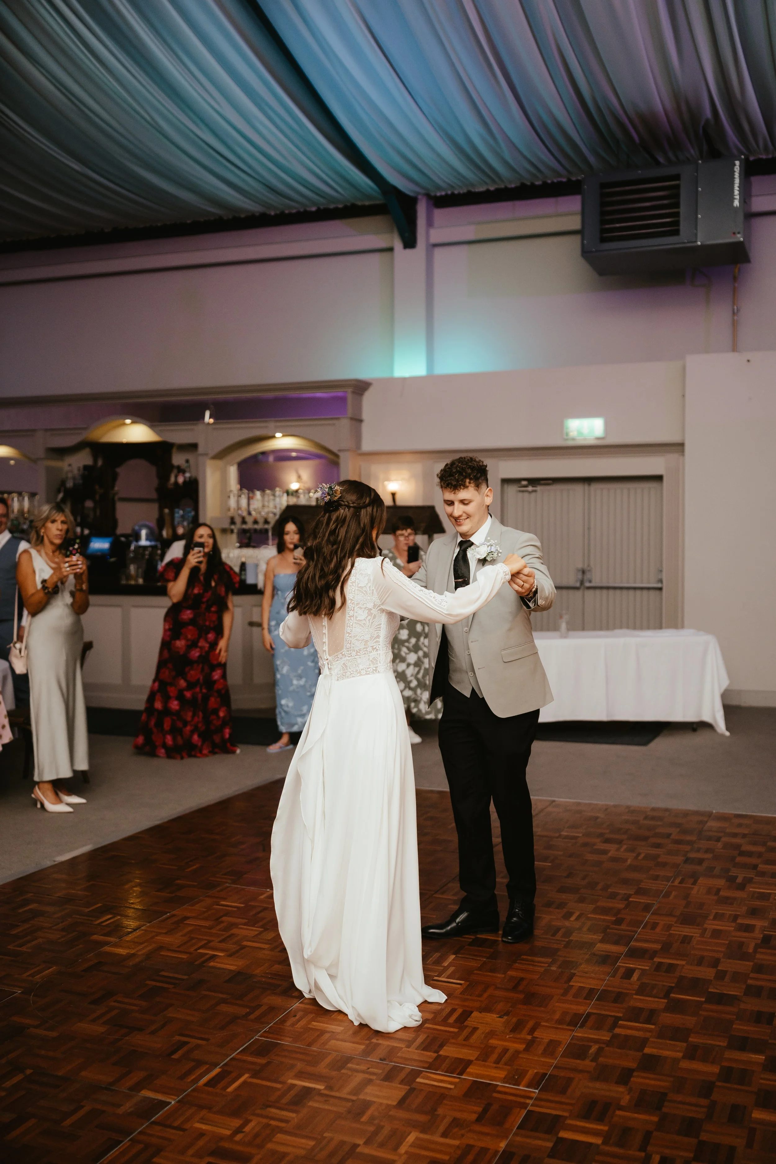 A bride and groom dance together on a wedding reception dance floor, surrounded by guests watching and taking photos in a decorated indoor venue.