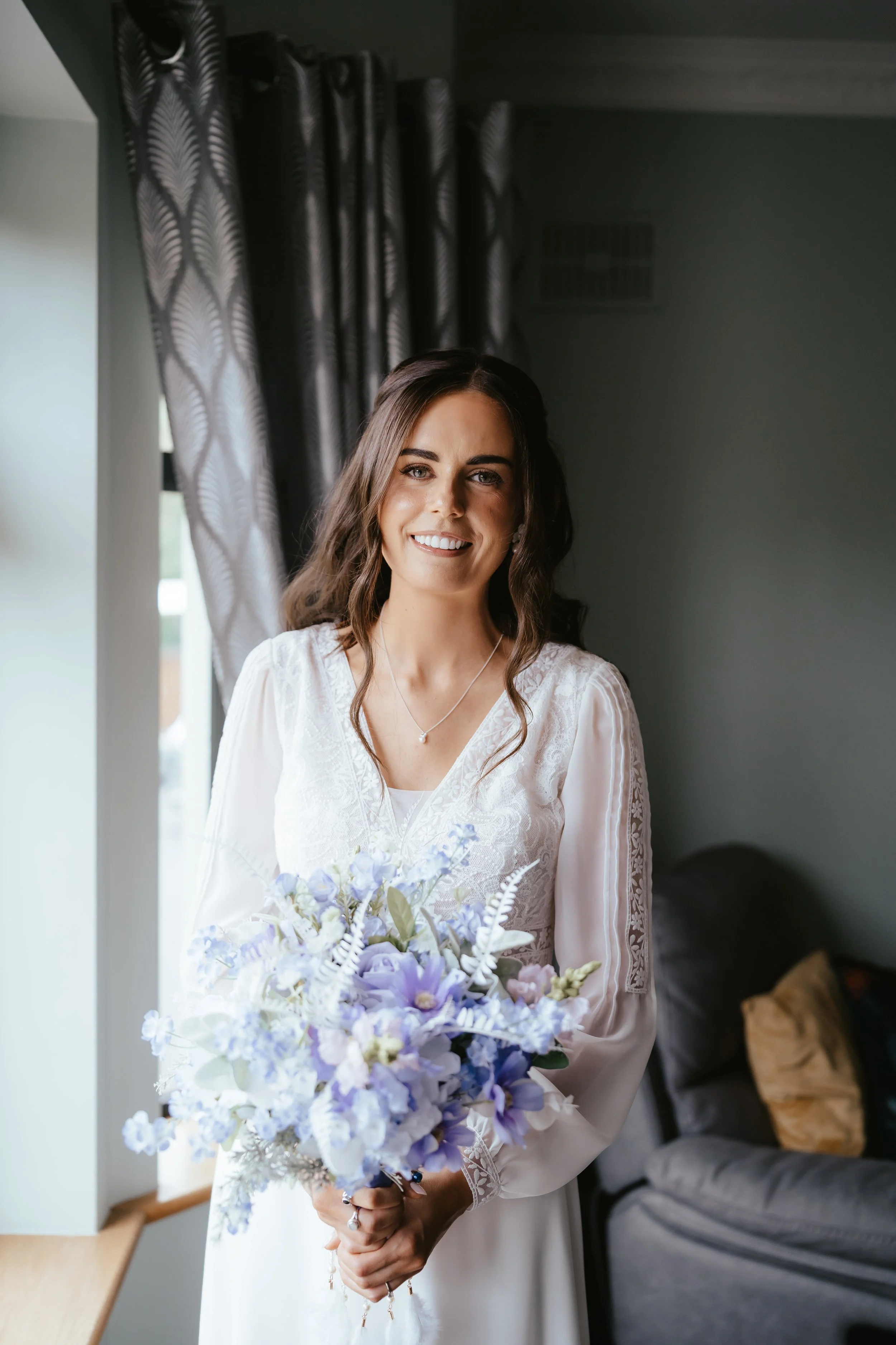 A young woman with long brown hair smiling, holding a bouquet of light purple and white flowers, in a room with dark curtains and a grey sofa.