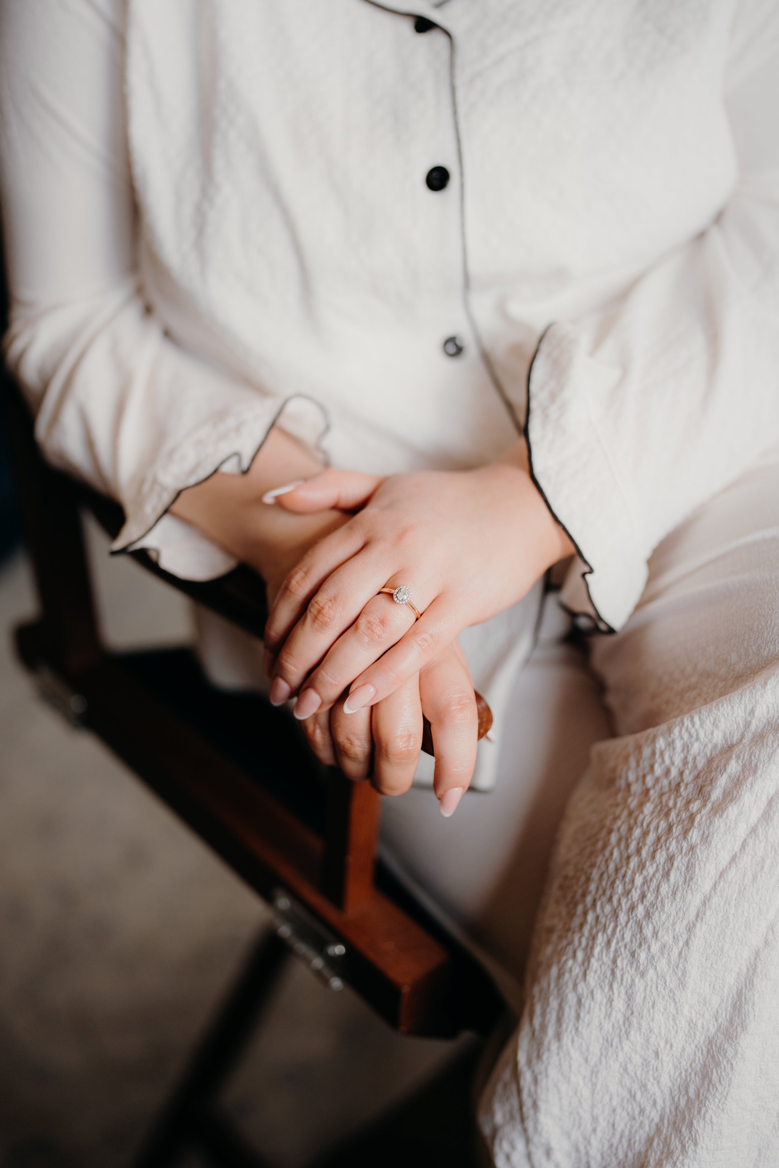 Close-up of a woman wearing a beige, textured shirt with black piping, sitting on a wooden chair with black metal legs, showing a diamond engagement ring on her left hand.