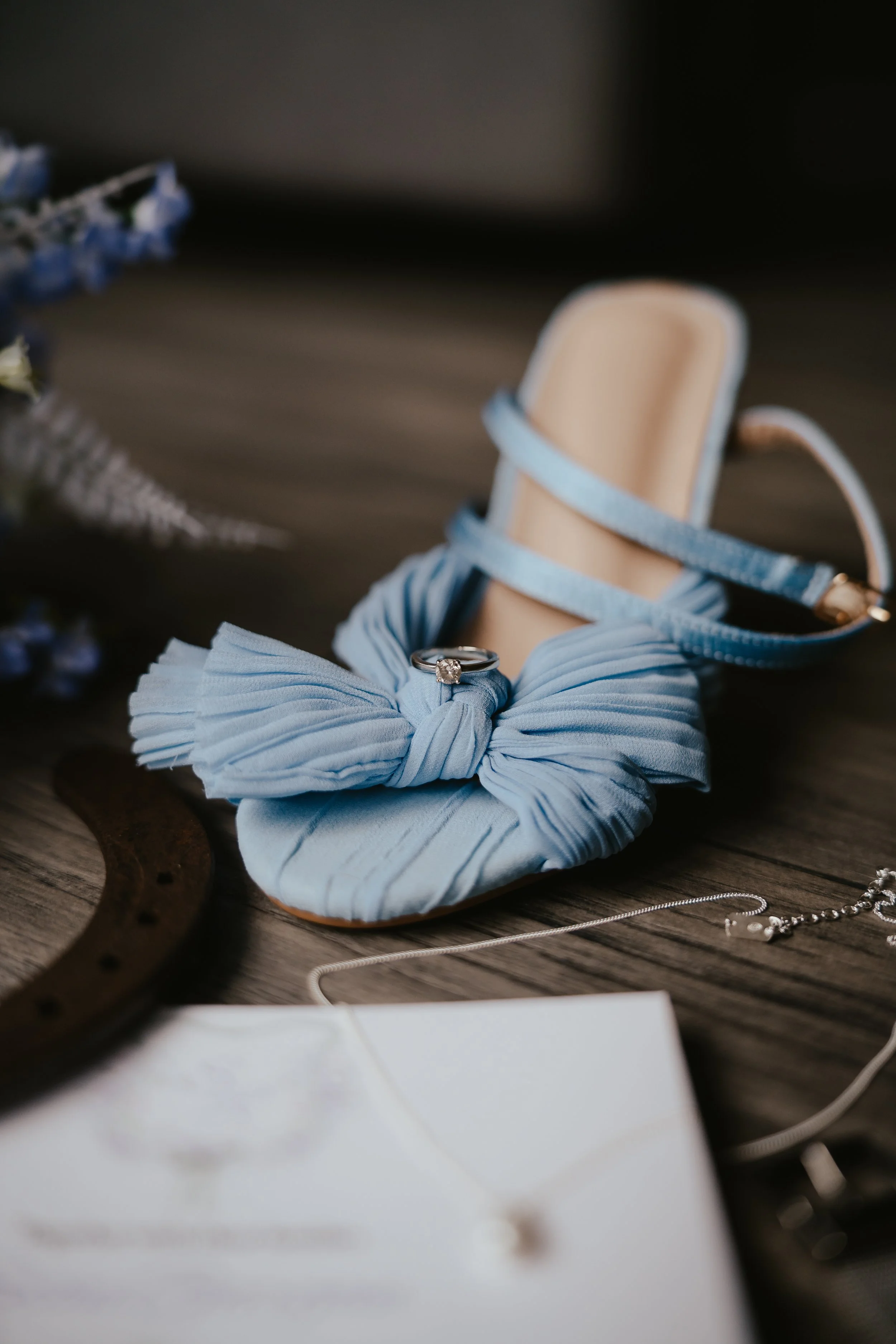 A blue fabric flower shoe with an engagement ring on top, along with a necklace, a wooden item, and some flowers in the background.