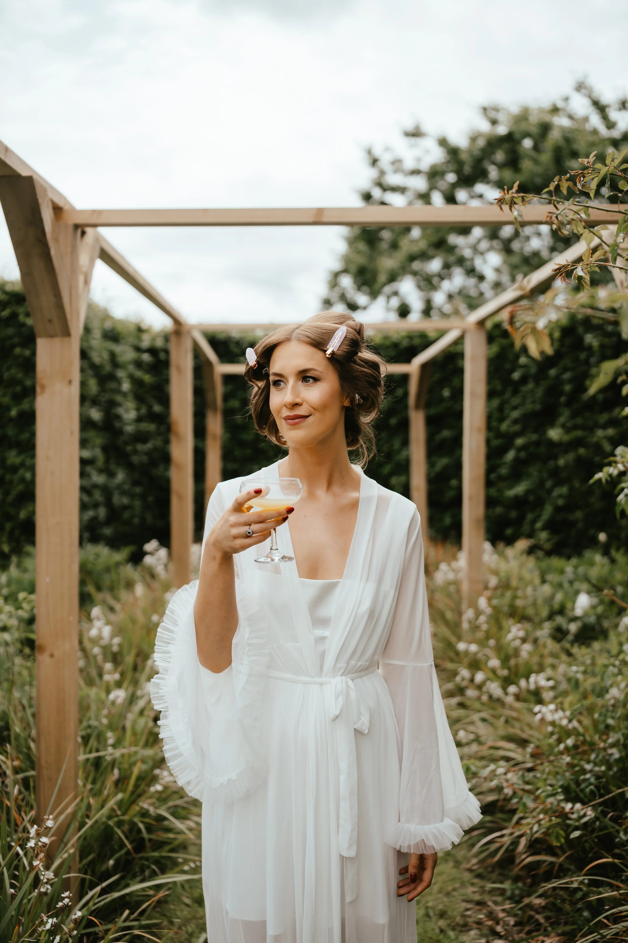 A woman in a white gown holds a cocktail glass while standing outdoors in a garden with a wooden structure and greenery.