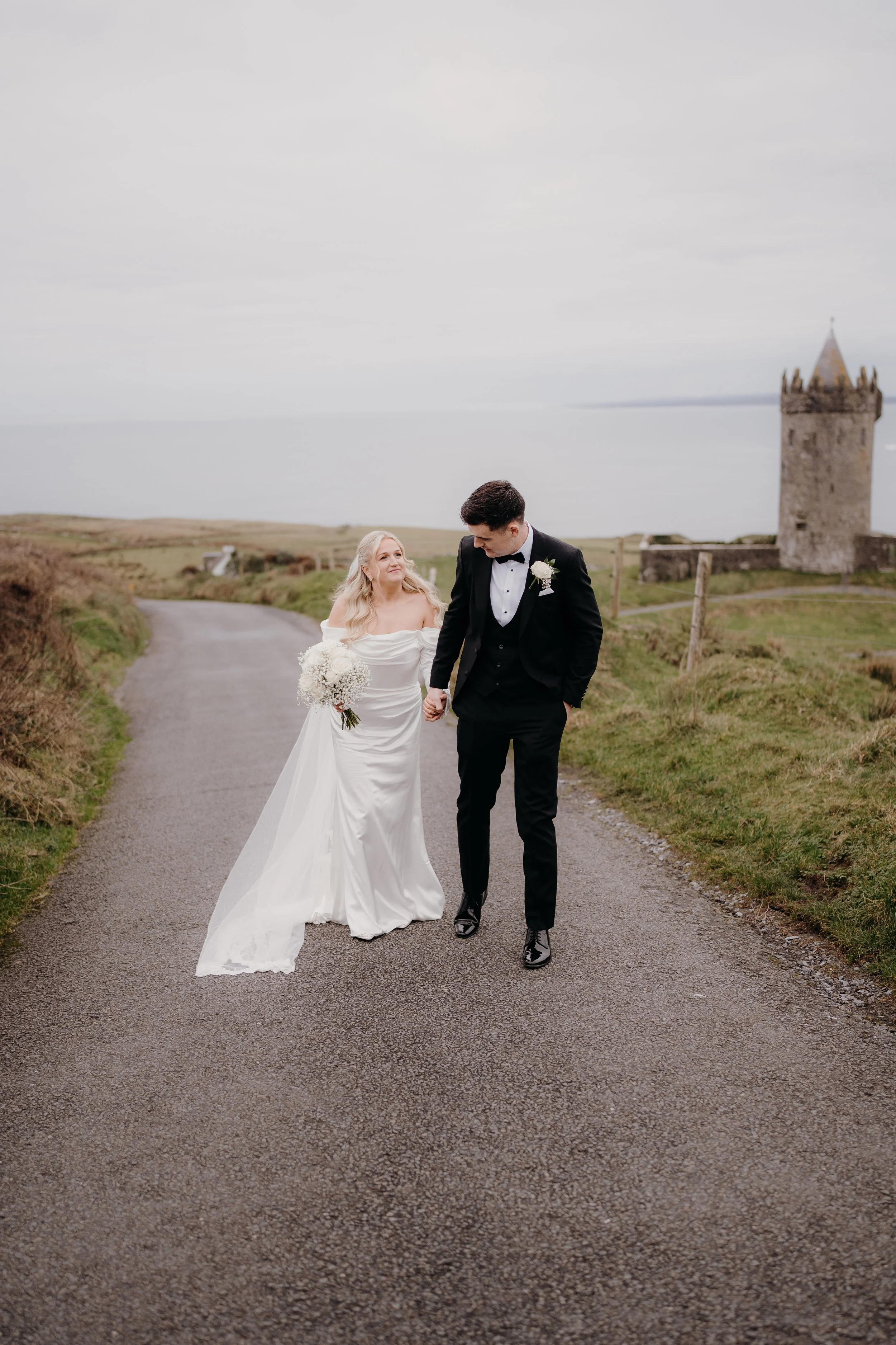 A bride and groom walking hand-in-hand on a rural road near a castle, with the bride in a white wedding dress holding a bouquet and the groom in a black tuxedo.