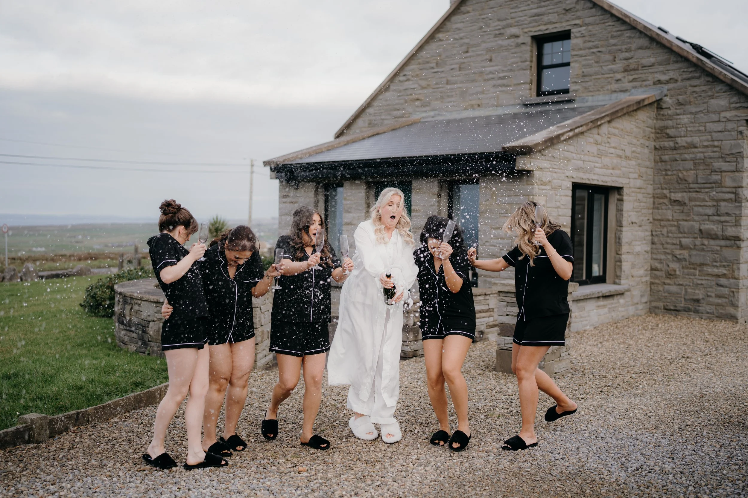 A group of women in black pajamas celebrating outdoors with champagne, some spraying champagne and celebrating, in front of a stone house on a cloudy day.