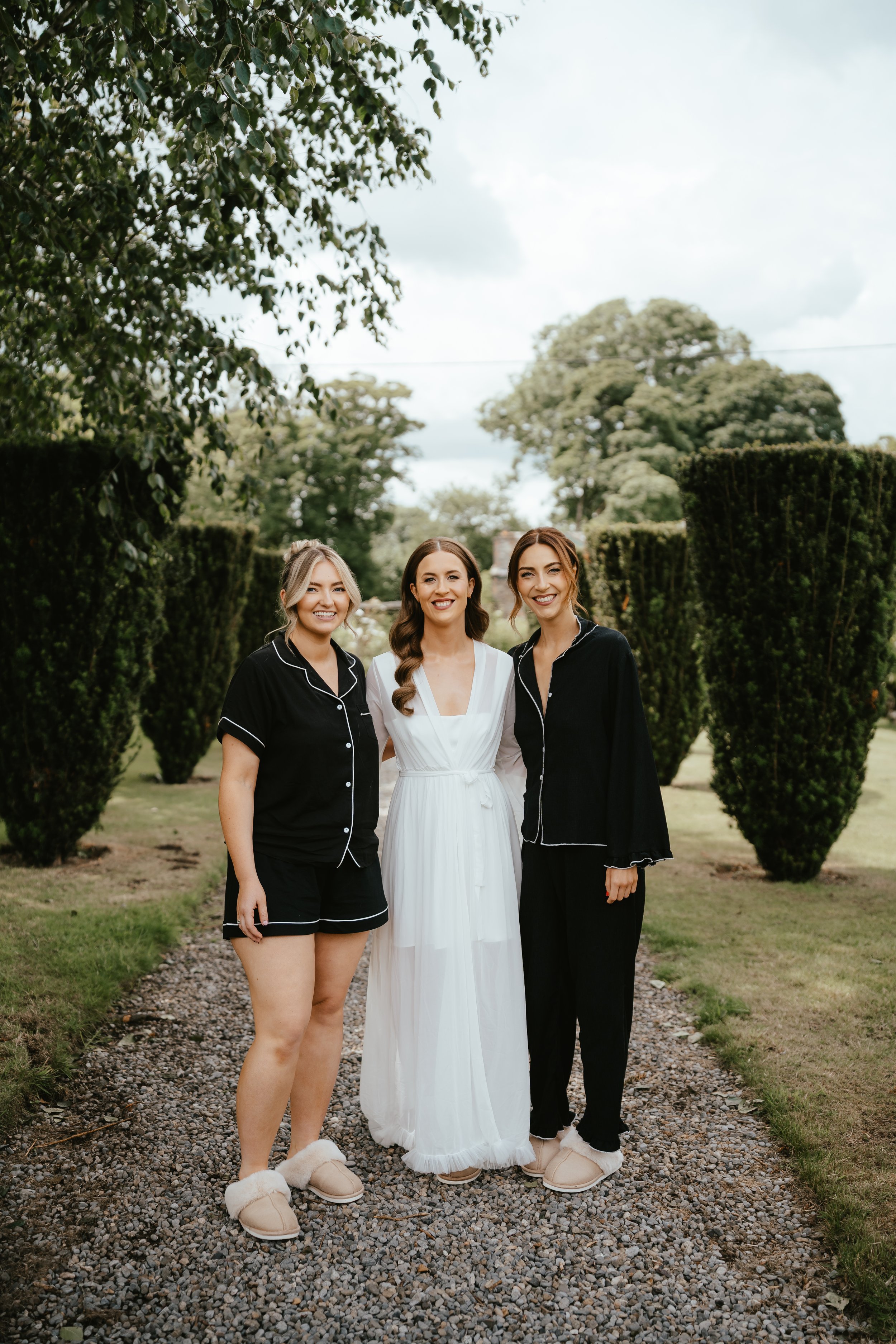 Three women standing outdoors on a gravel path, smiling, with trees and bushes in the background. The woman in the middle is wearing a white dress, and the other two women are dressed in black pajamas, all wearing slippers.