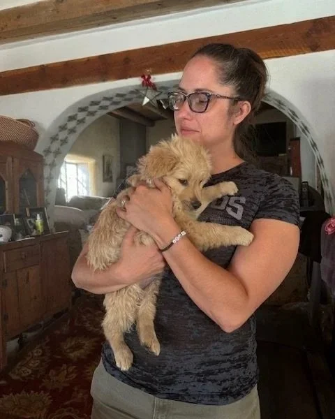 A woman wearing glasses holding a small golden retriever puppy inside a home with rustic decor.