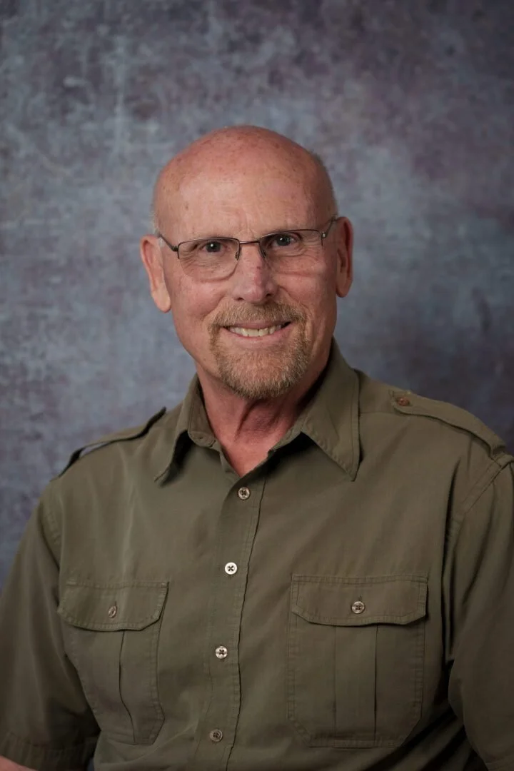 A smiling man with glasses, a beard, and a bald head, wearing an olive green button-up shirt, posing against a textured background.