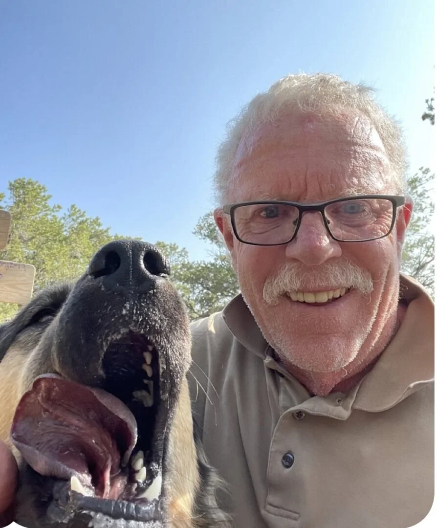 An elderly man with glasses smiling next to a yawning German Shepherd dog during the daytime with trees and a blue sky in the background.