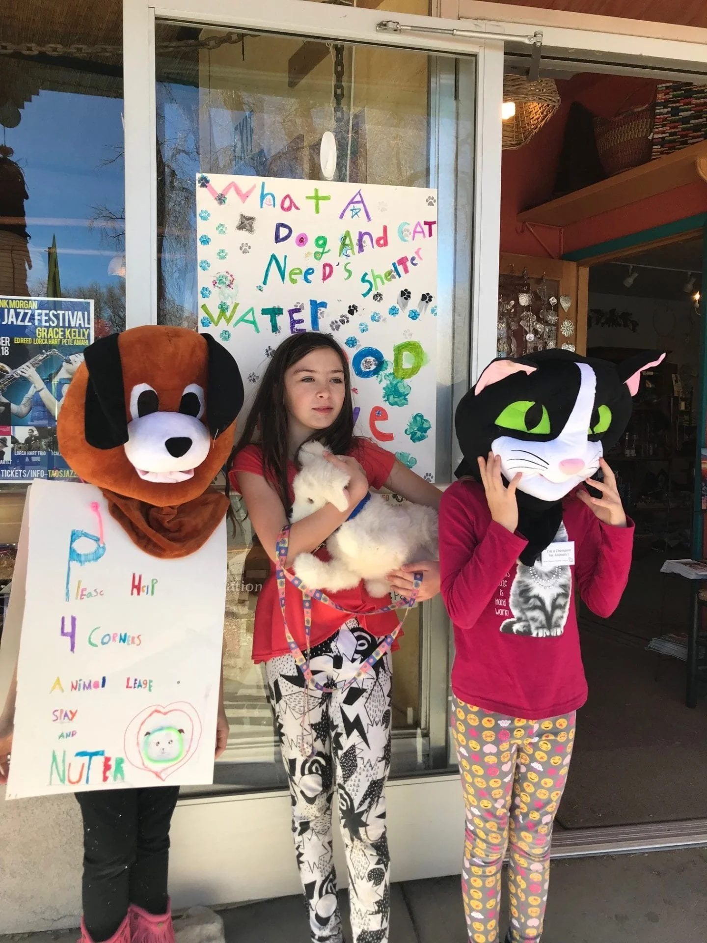 Two children wearing animal masks and holding a puppy and a plush toy, standing next to a girl with a sign about animal shelter needs outside of a store with a colorful poster in the background.