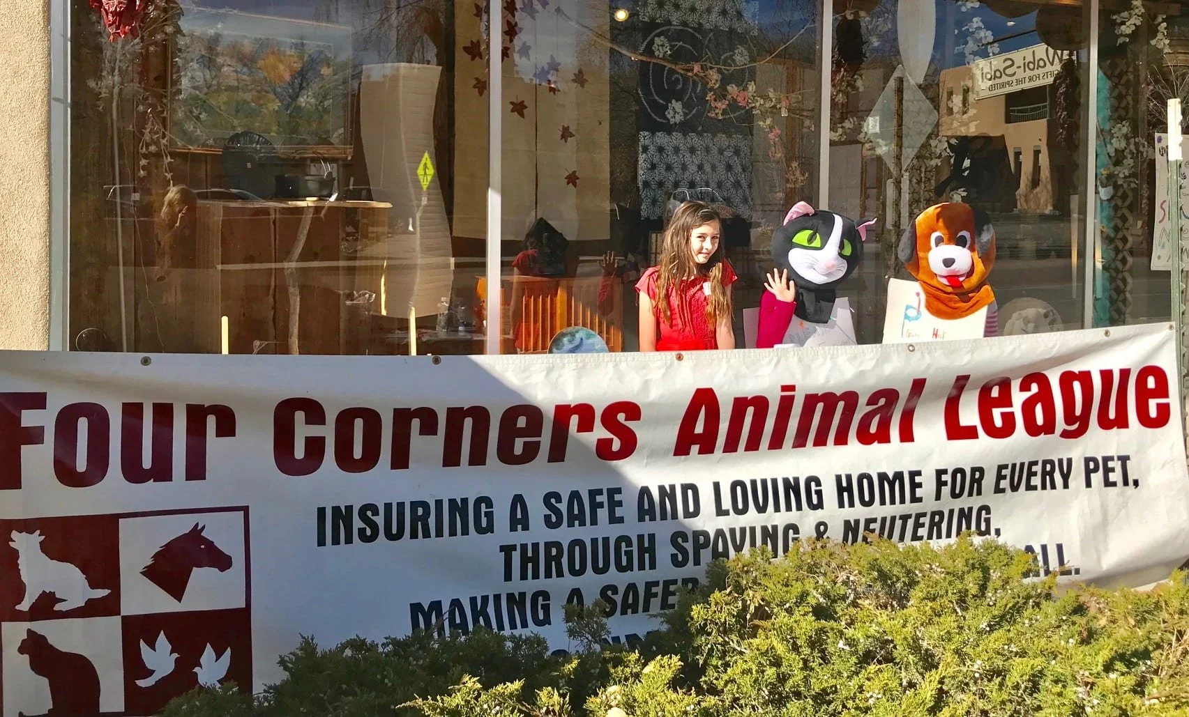 A storefront window display with a girl in a red dress and three people in animal costumes, including a black and white cat, a brown and white dog, and a white kid. A banner in front promotes the Four Corners Animal League, advocating for safe and lo